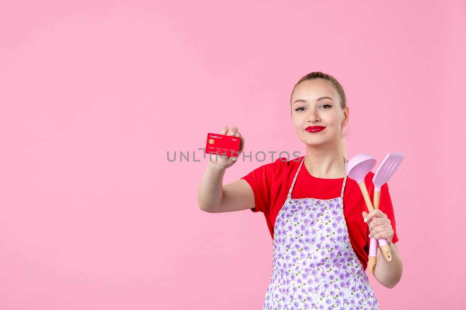 front view young housewife in cape holding spoons and red bank card on pink background profession occupation duty money uniform job cutlery worker horizontal