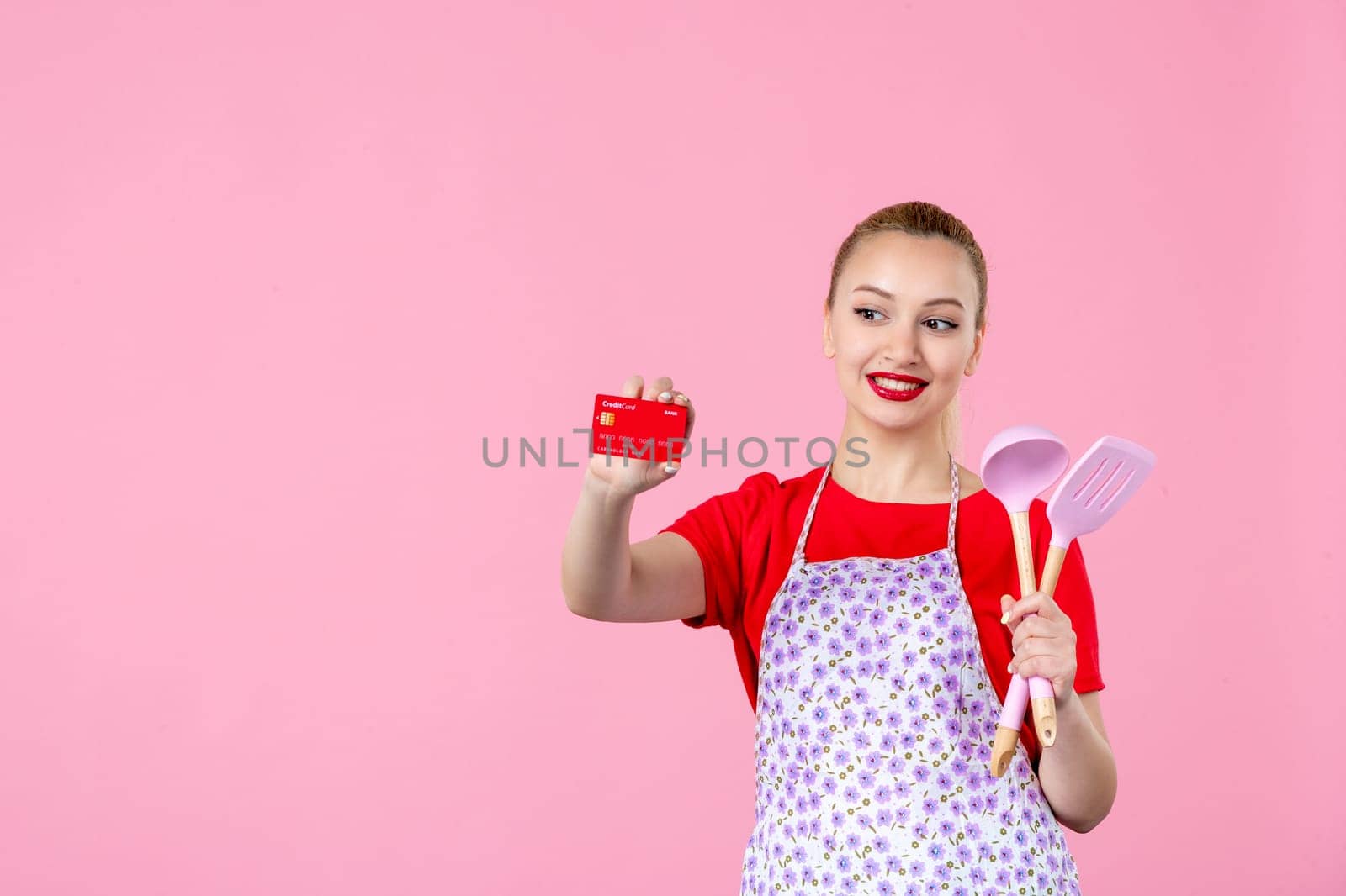 front view young housewife in cape holding spoons and red bank card on pink background profession occupation duty money uniform job cutlery worker horizontal wife