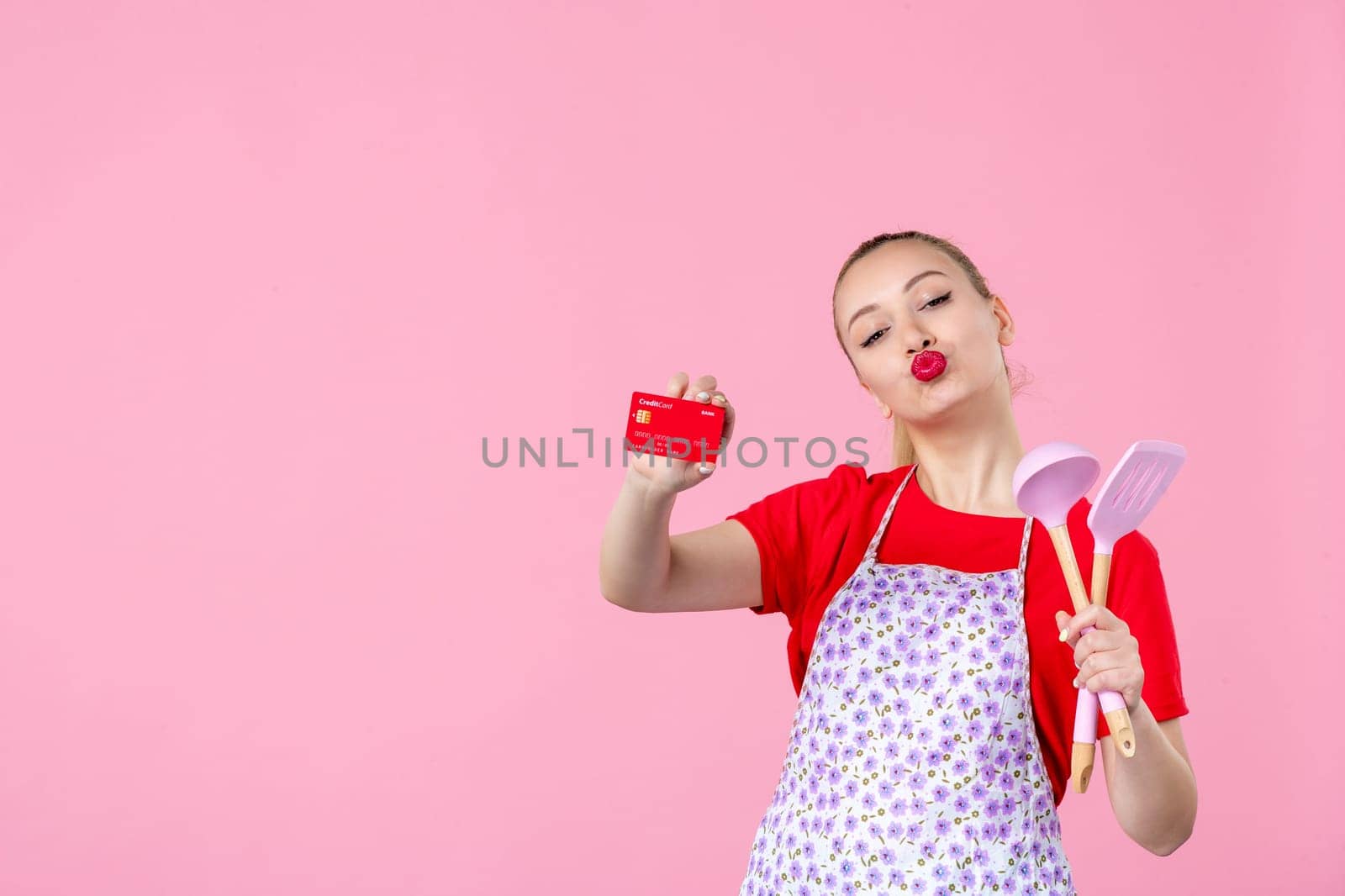 front view young housewife in cape holding spoons and red bank card on pink background profession occupation duty money uniform job cutlery worker wife by Kamran