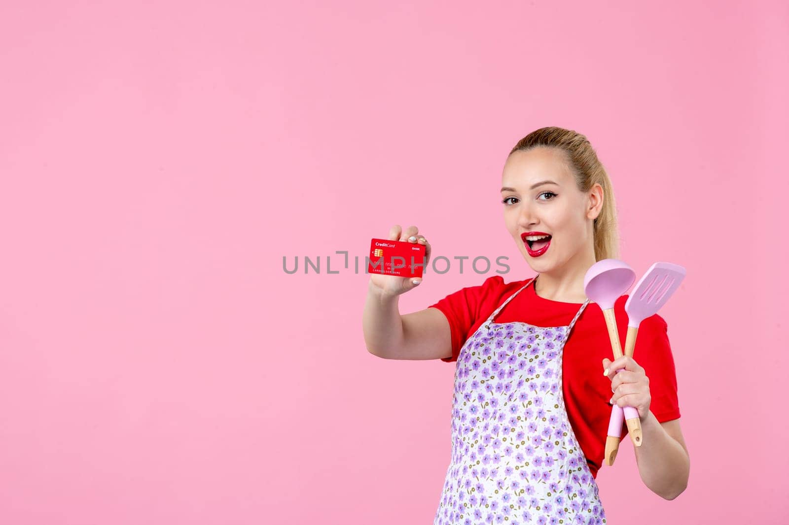 front view young housewife in cape holding spoons and red bank card on pink background profession occupation duty money uniform job worker horizontal wife by Kamran
