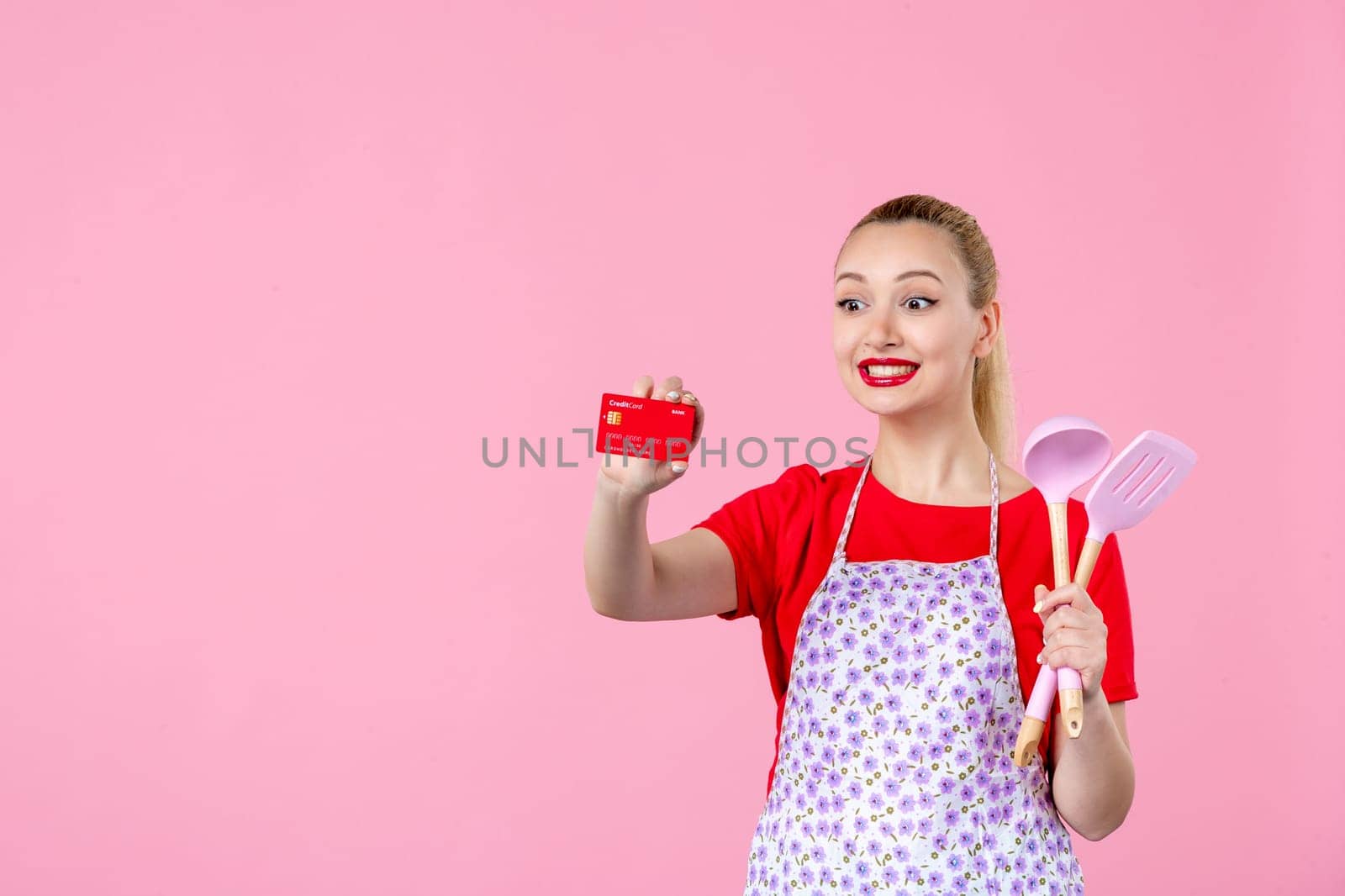 front view young housewife in cape holding spoons and red bank card on pink background profession occupation duty money wife uniform job cutlery worker horizontal