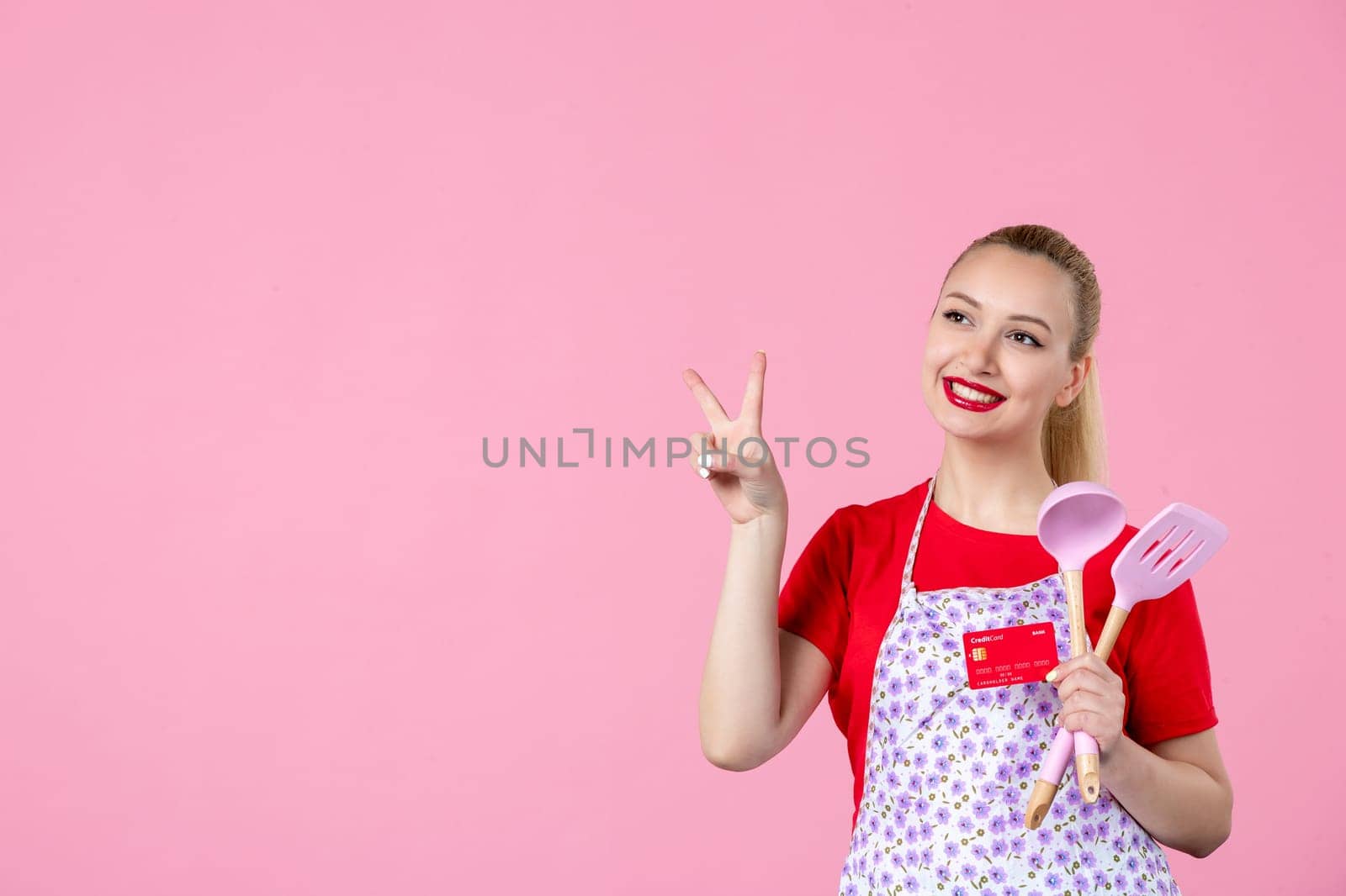 front view young housewife in cape holding spoons and red bank card on pink background worker wife duty profession horizontal occupation uniform job