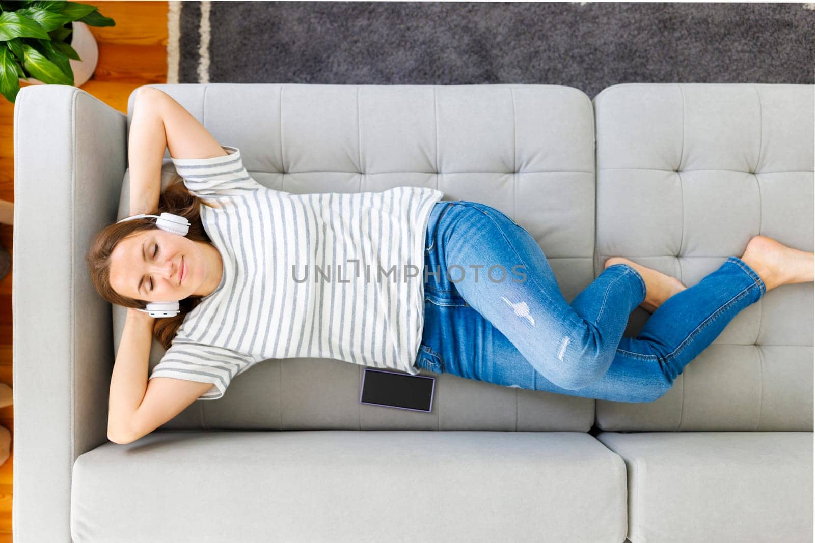 Peaceful woman in modern wireless headphones laying, relaxing on a comfortable couch, listening to music. Happy calm woman wearing headphones relaxing on a comfortable sofa, enjoy quality sound