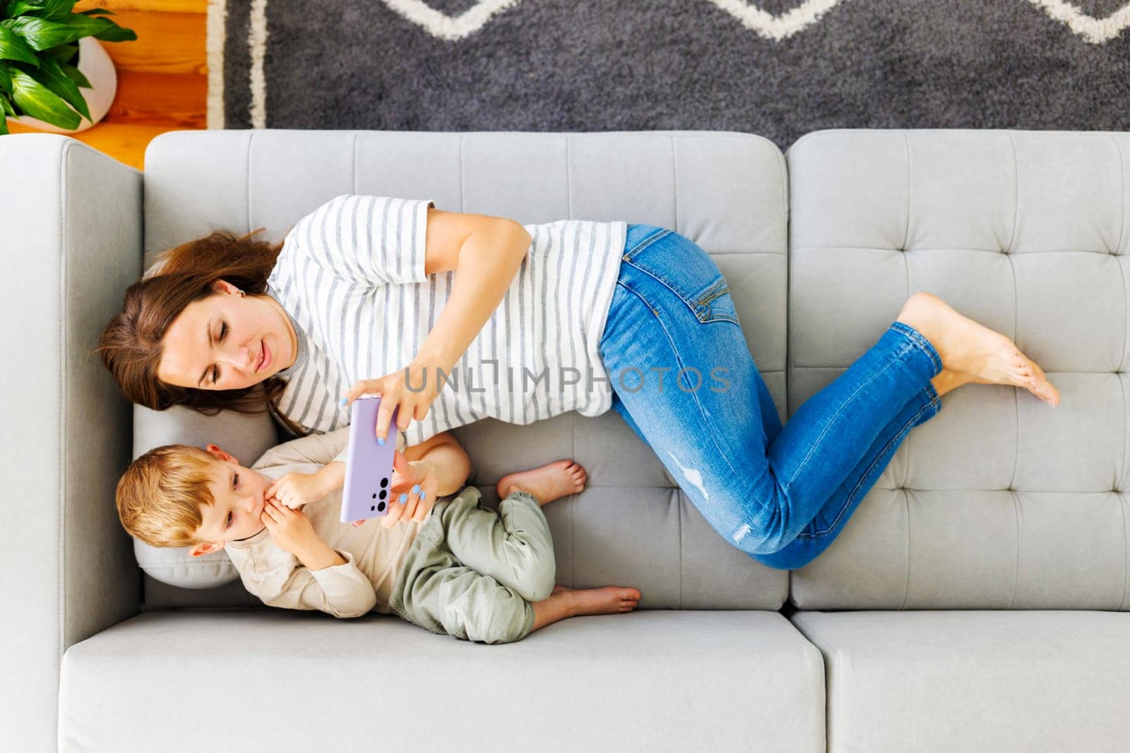 Woman and her little son lying on cozy sofa with smartphone, above view. Family spend leisure together at home using smartphone, play games, watch video, take selfies