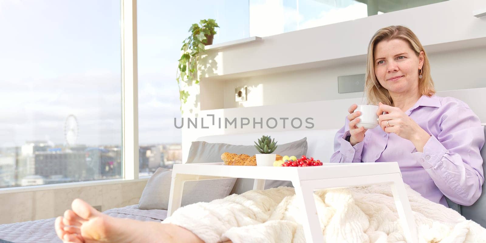 Happy middle aged, beautiful woman having breakfast, relaxing in hotel bed. Woman having breakfast sitting on her bed in luxury apartments. enjoying lazy morning lifestyle