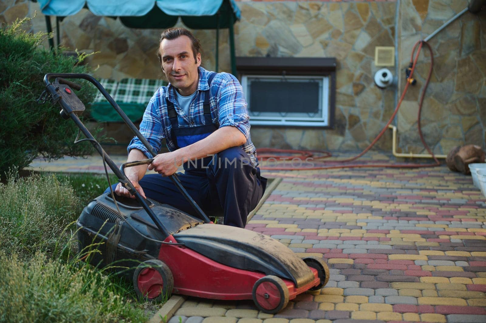 Portrait of an attractive Caucasian man, worker gardener, professional landscaper 40 years old, smiling looking at camera while replacing a filter on the electric lawn mower in the garden backyard.