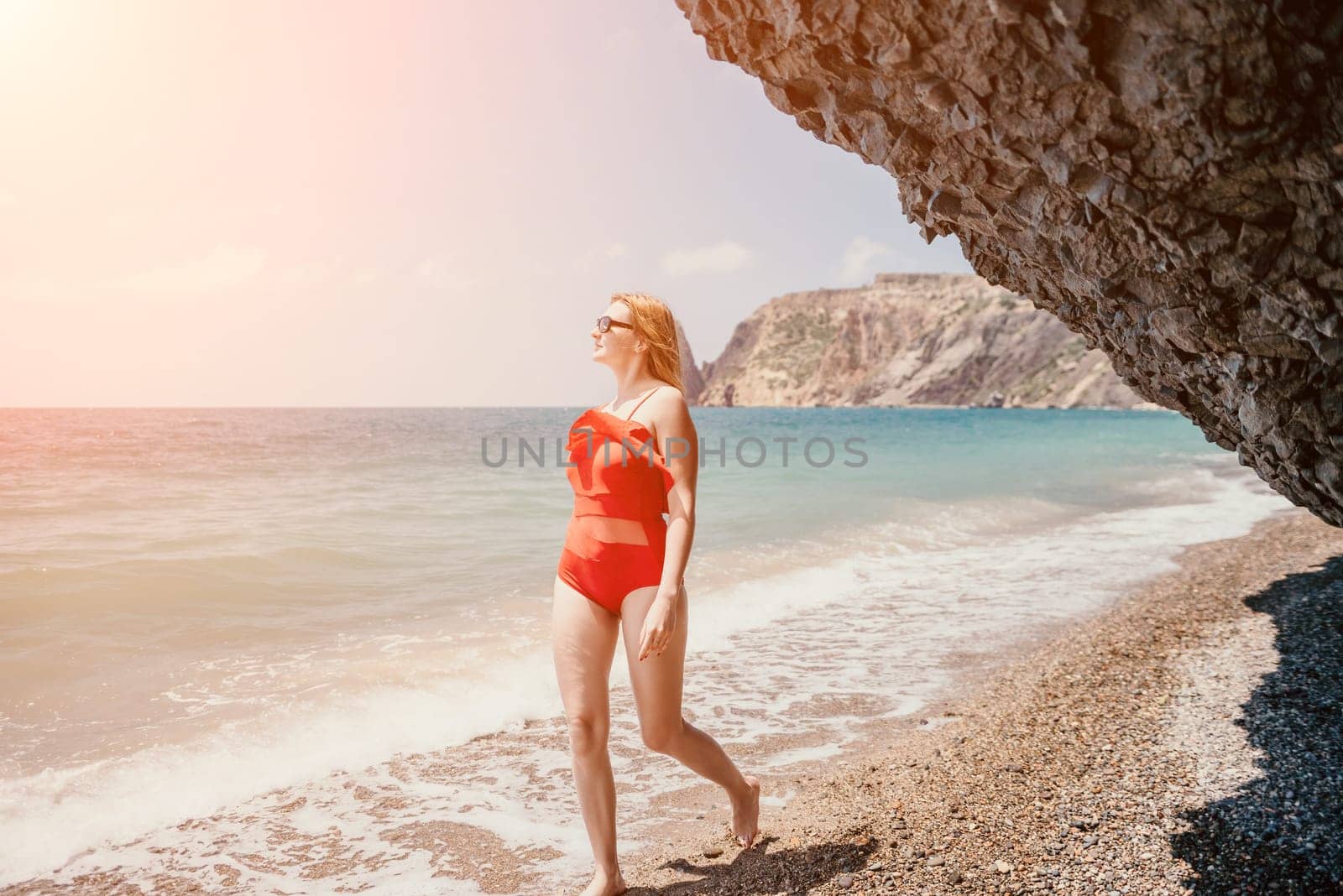 Young woman in red bikini on Beach. Blonde in sunglasses on pebble beach enjoying sun. Happy lady in one piece red swimsuit relaxing and sunbathing by turquoise sea ocean on hot summer day. Close up,