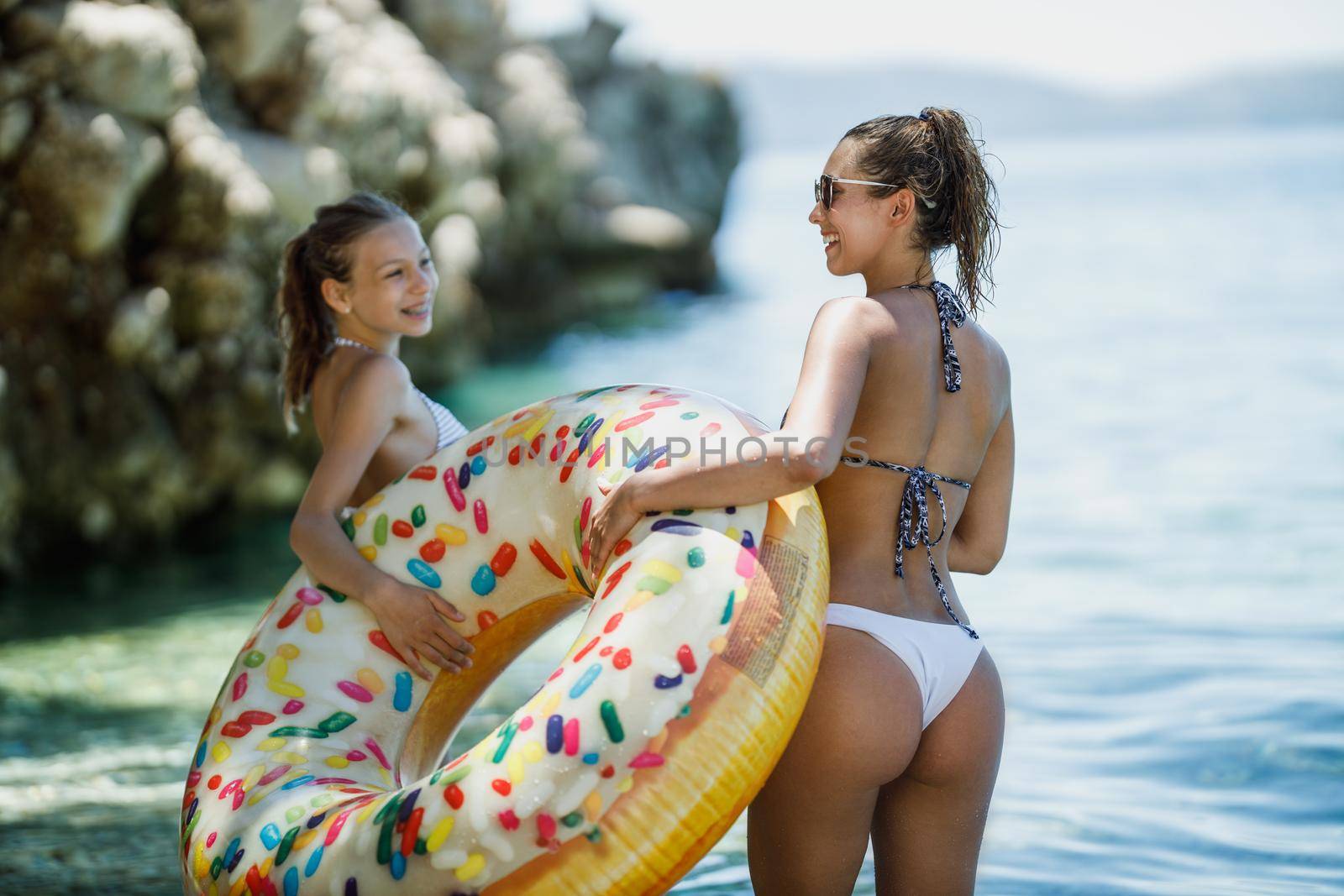 A beautiful teenage girl is enjoying with her young mother on the beach. They are having fun and bringing an inflatable rubber circle in the water to swim.