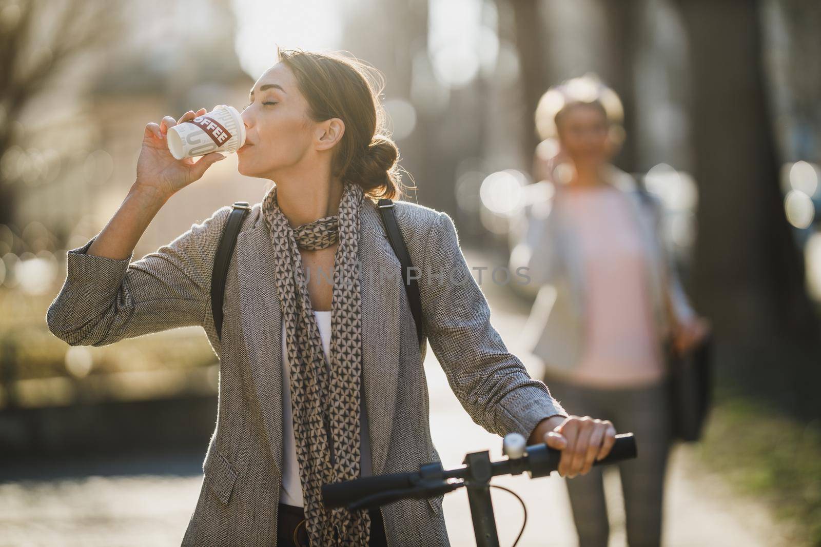 A young businesswoman drinking coffee while traveling with an electric scooter through the city.