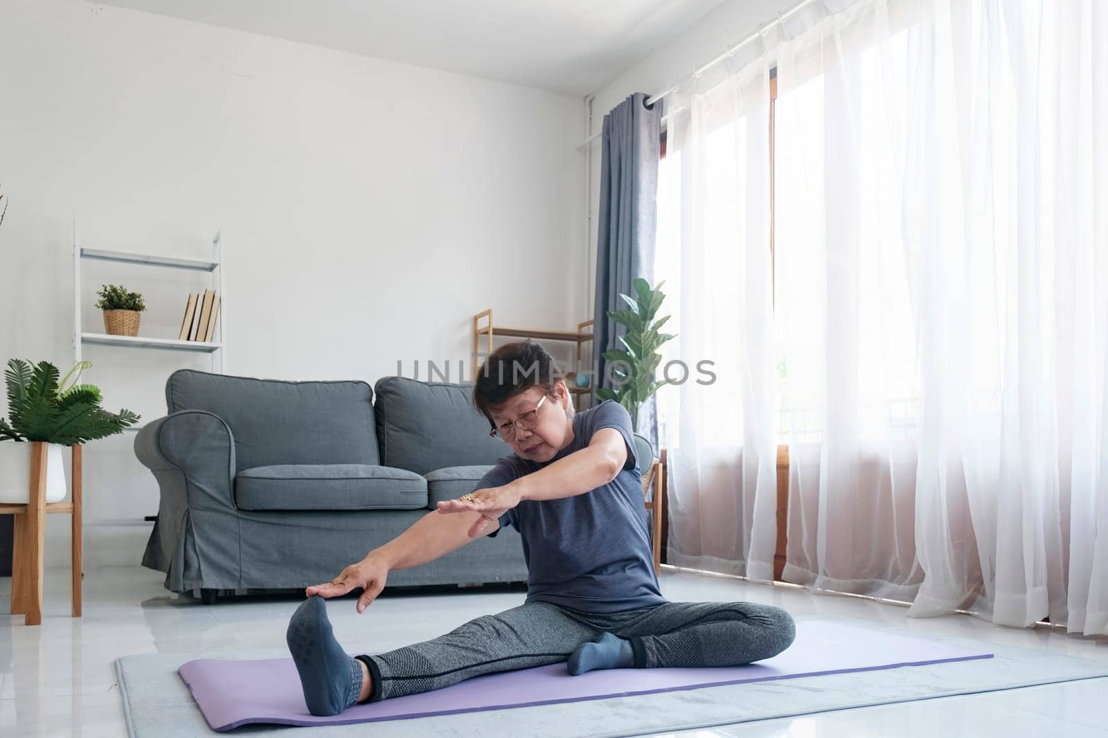 Active mature woman doing stretching exercise in living room at home. Fit lady stretching arms and back while sitting on yoga mat.