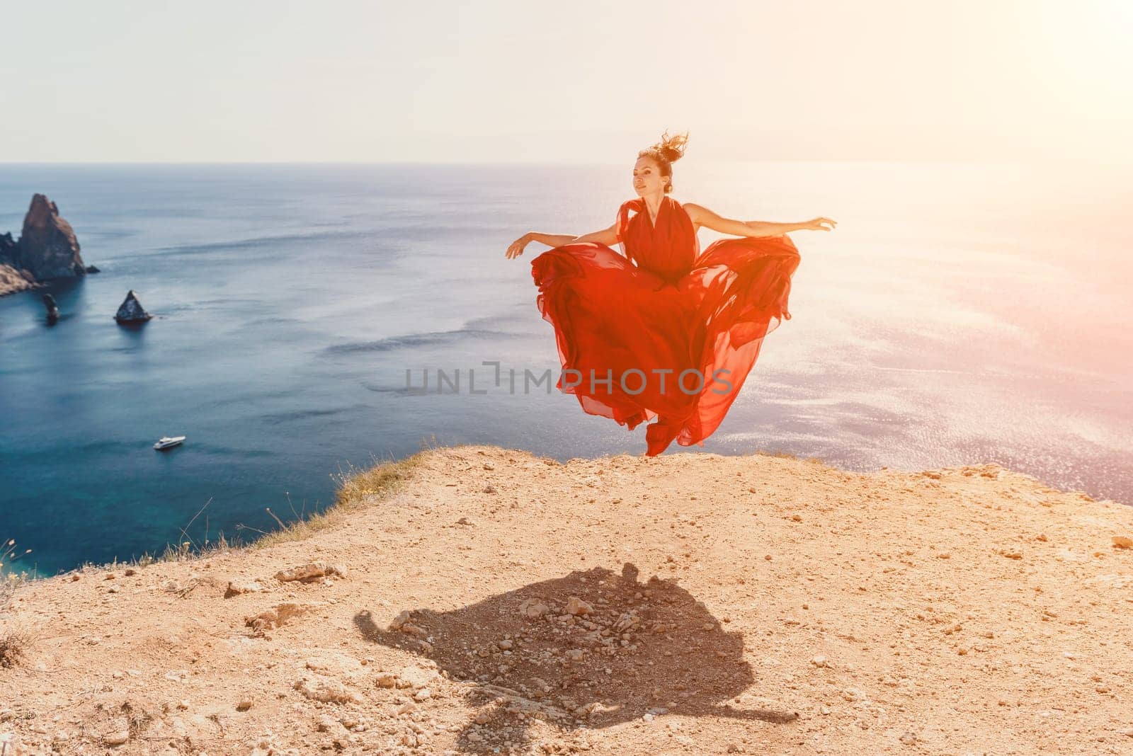 Woman in red dress on sea. Side view a Young beautiful sensual woman in a red long dress posing on a rock high above the sea on sunset. Girl on the nature on blue sky background. Fashion photo. by panophotograph
