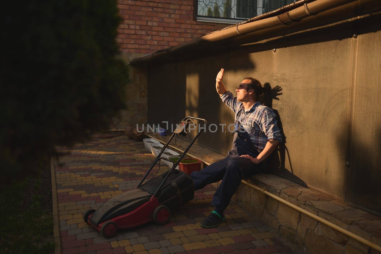 Portrait of a worker looking at sunset after finishing hard working day in the garden, holding his hand over his face, sitting outdoors near a lawn mower, relaxing. People. Workman. Labor. Lifestyle