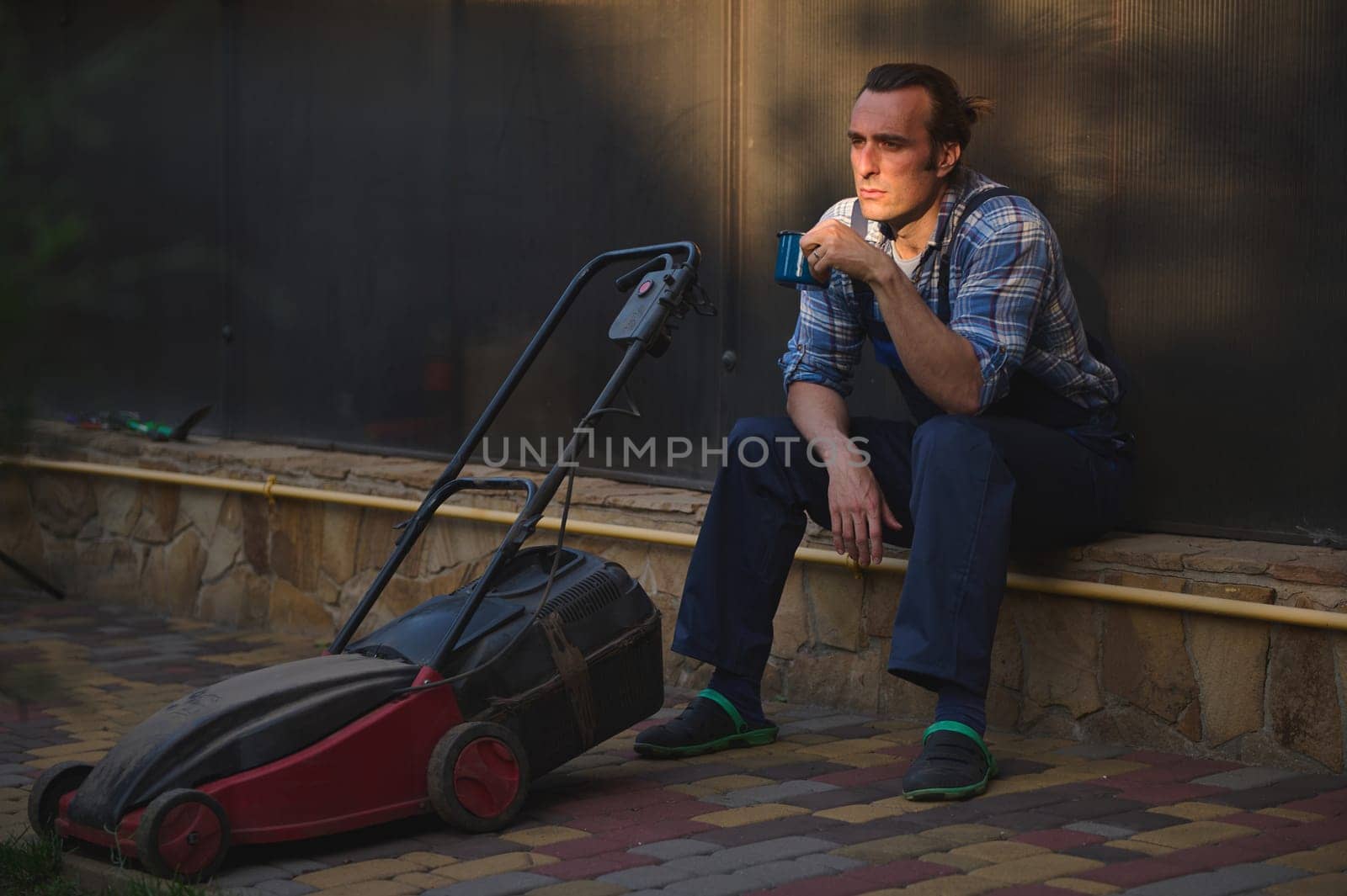 Sunset sun rays fall on face of a gardener with lawn mower, overworked after hard day's work in the garden, relaxing with a cup of coffee. Landscaping hobby and industry. People. Labour. Recreation