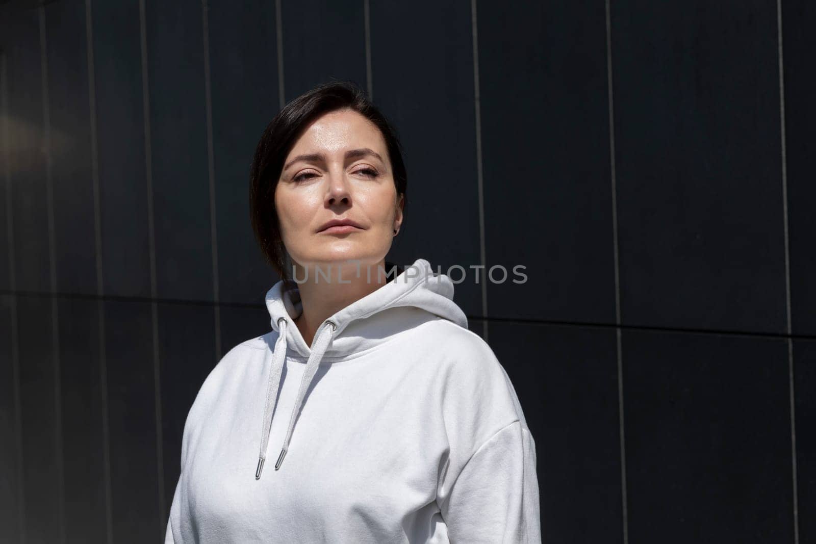 Portrait Smiling Mature Brunette Woman in White Hoodie Looking Away, Dark Background Wall. Sportive Relaxed 40 Yo Beautiful Caucasian Woman in City. Horizontal Plane. High quality photo