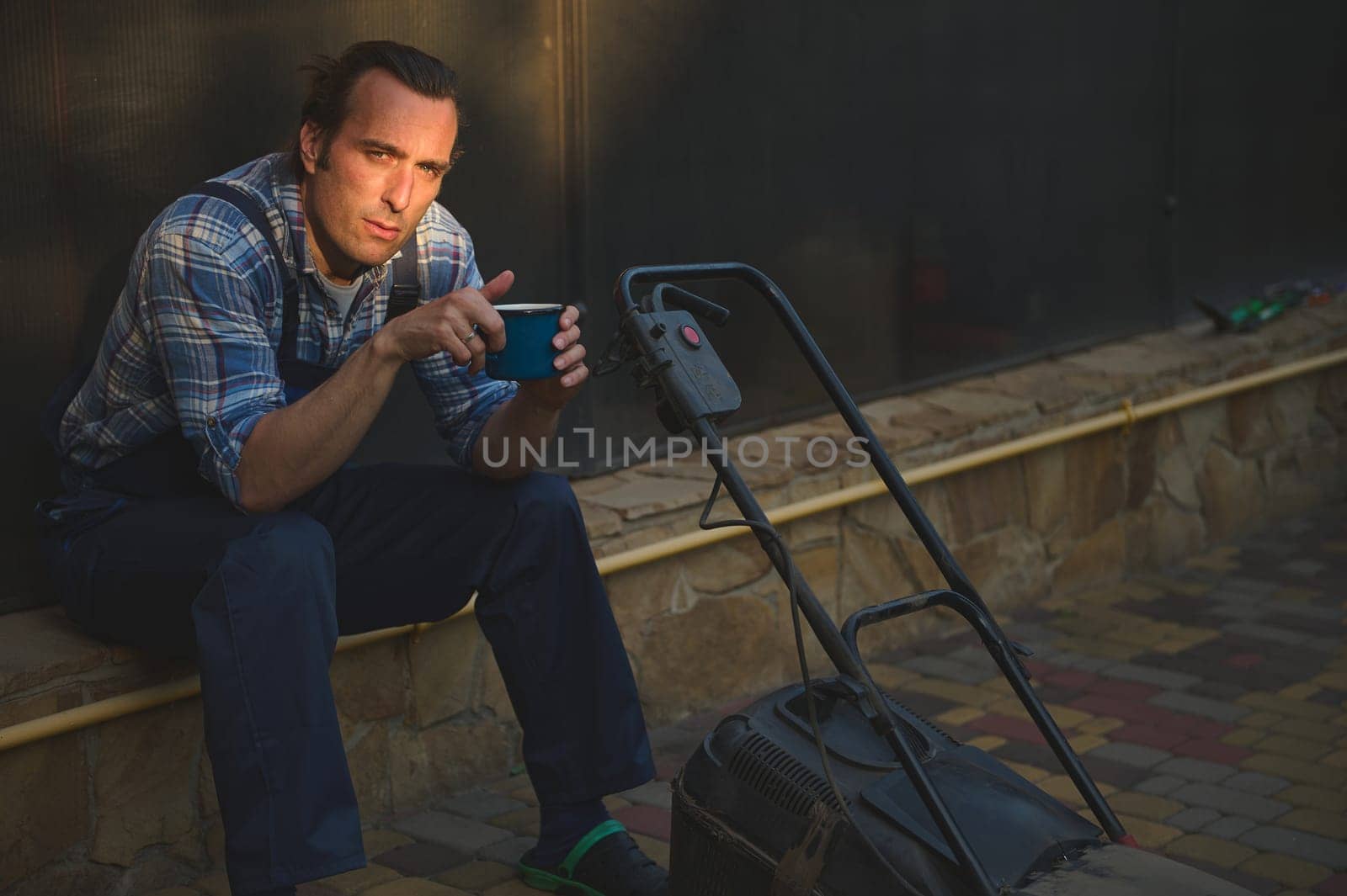 Attractive gardener man in blue work jumpsuit, looking at camera, sitting near a lawn mower with cup of coffee in hands, relaxing after hard working day in the garden. Sunset sun rays fall on his face