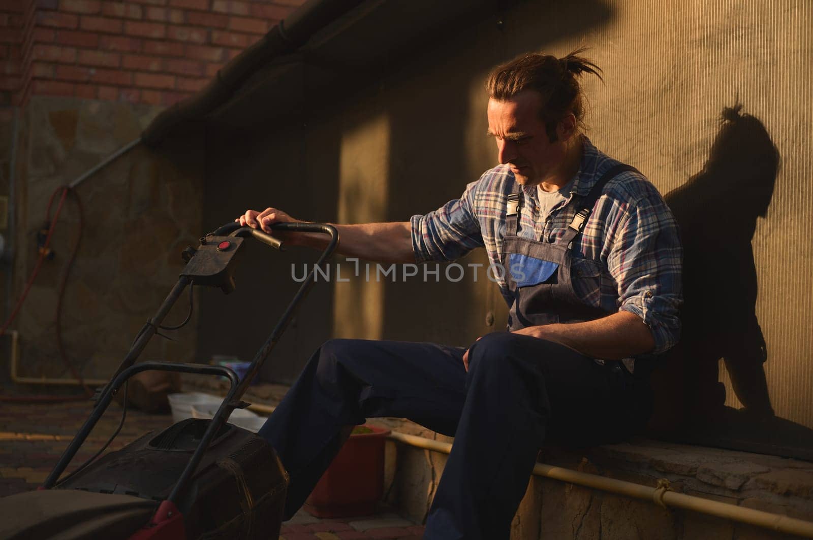 Portrait of overworked gardener man, homeowner, landscaper in blue gardening overalls and checkered shirt, with a lawn mower in the garden at sunset, relaxing after a hard day's work. People. Labour