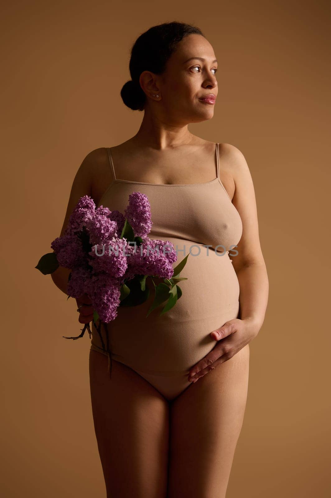 Portrait of a beautiful pregnant woman expecting baby, holding belly, looking aside, posing in lingerie with a bunch of purple lilacs, isolated beige studio background. Pregnancy and maternity concept