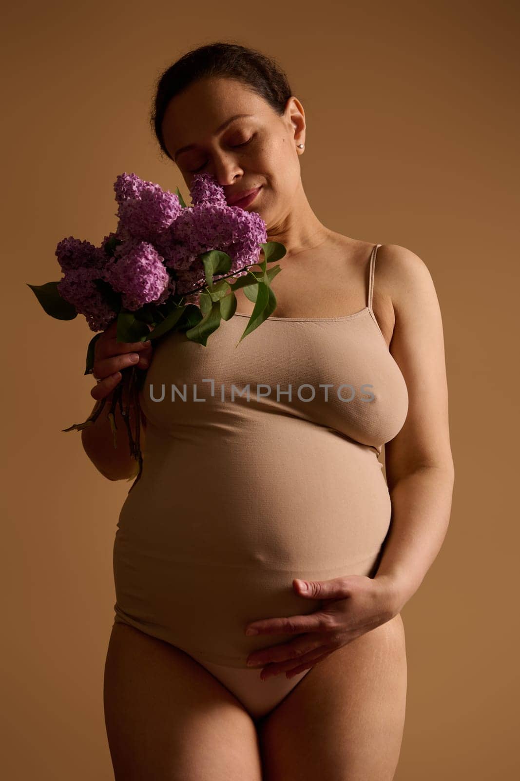 Studio portrait of a beautiful adult pregnant woman caressing her belly, holding a bunch of lilacs, isolated on beige by artgf