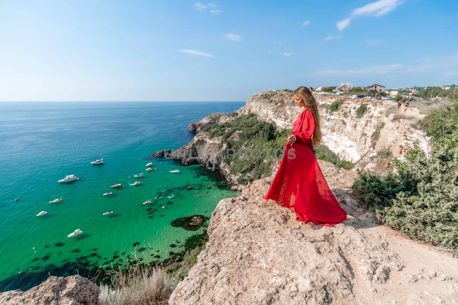 Red Dress Woman sea Cliff. A beautiful woman in a red dress and white swimsuit poses on a cliff overlooking the sea on a sunny day. Boats and yachts dot the background. by Matiunina
