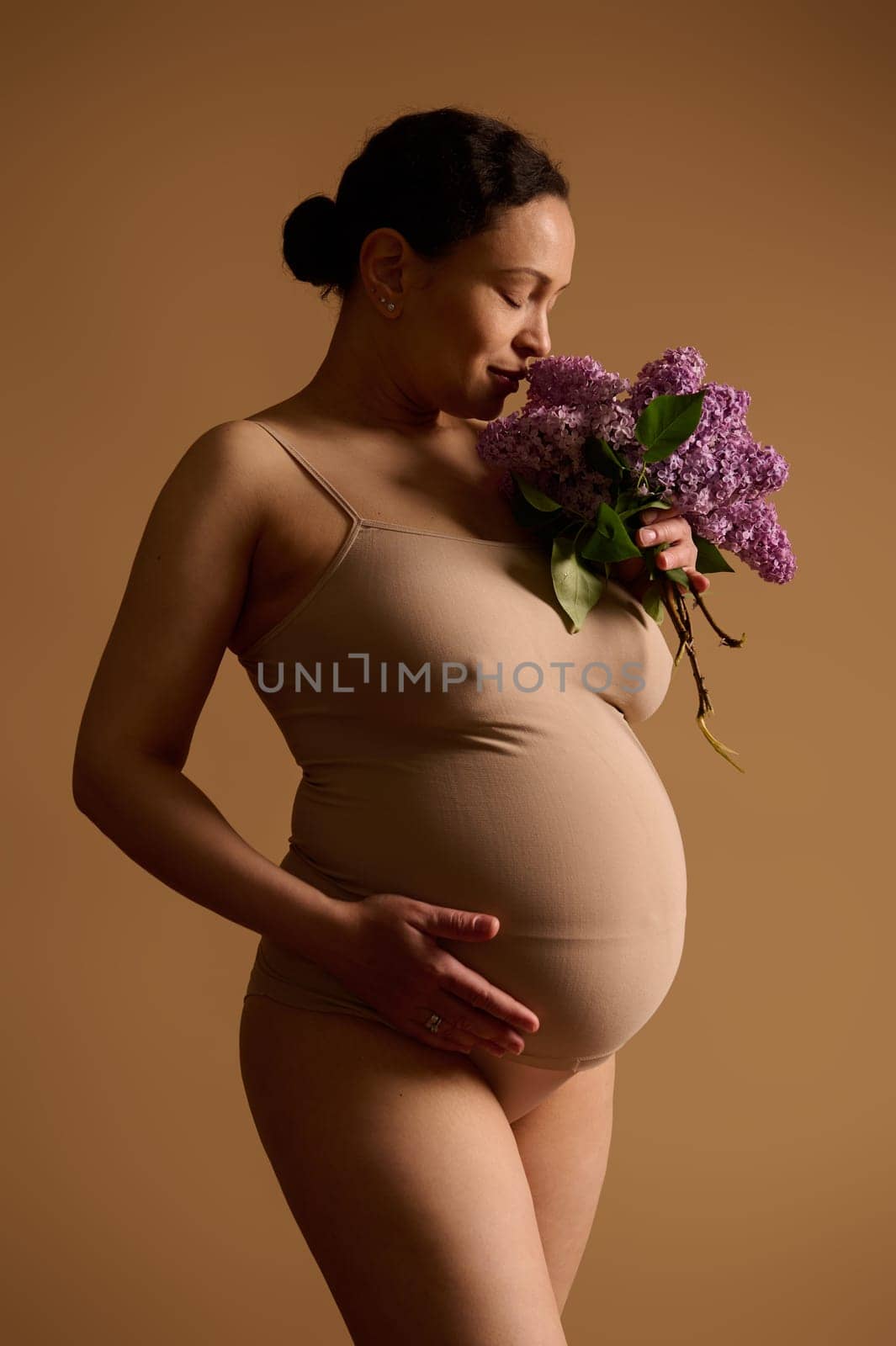 Beautiful pregnant woman in beige lingerie, holding hand on her big belly, smelling a bunch of blooming purple lilacs, studio background. Pregnancy, Gynecology, Human fertility. Women's health concept