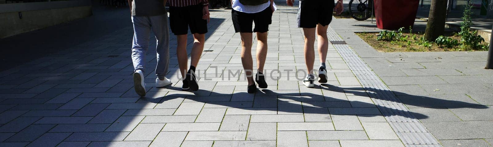 The legs of four male friends. They walk on a broad boulevard. Three of them have bare legs. Their shadows are visible on the paving stones