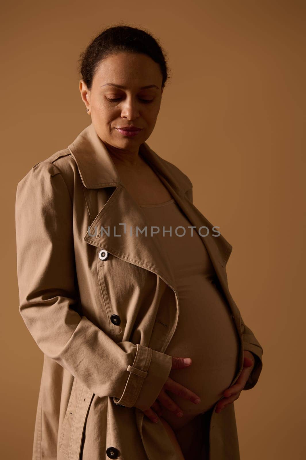 Portrait of African adult pregnant woman in trendy trench coat, holding hands on her belly, isolated on cream background by artgf