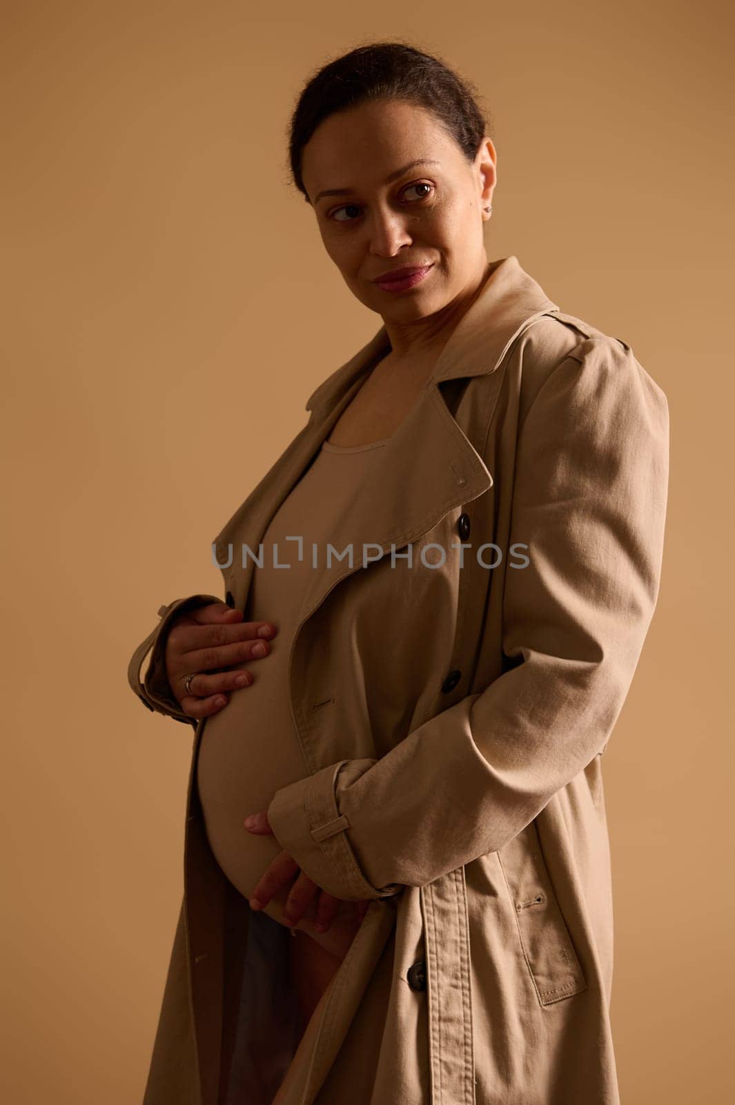 Gorgeous gravid woman dreamily looking away, touching her pregnant belly, feeling baby kicks, isolated beige background by artgf