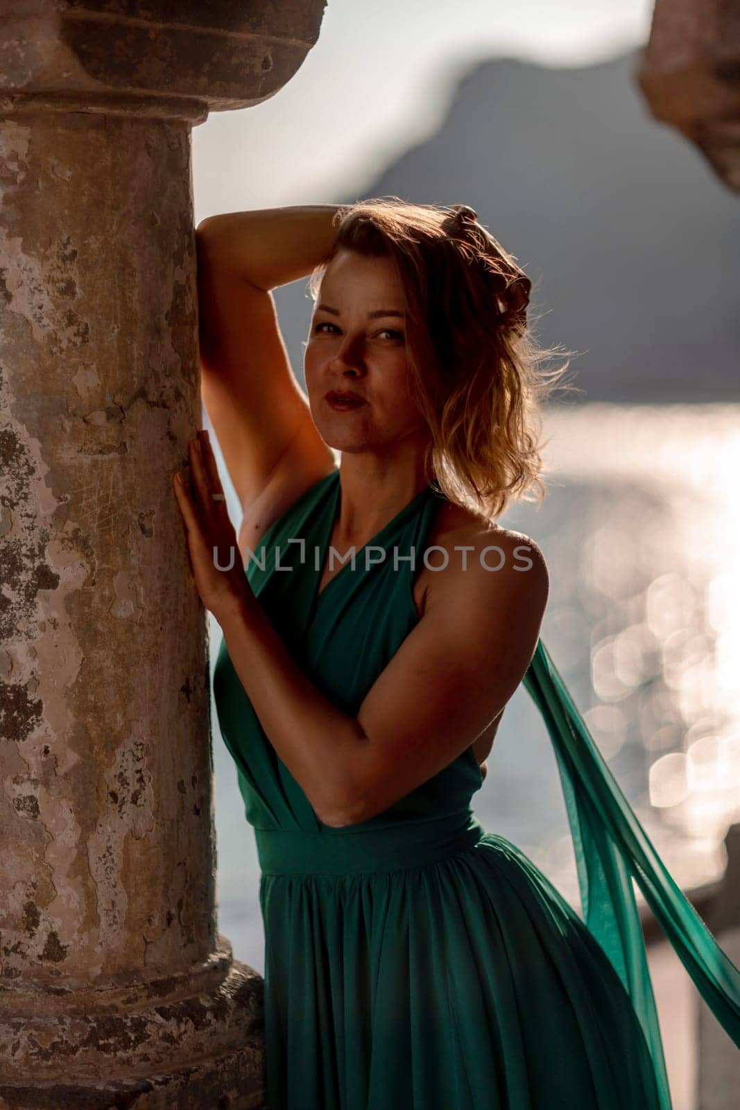 Happy blonde in a long mint dress posing against the backdrop of the sea in an old building with columns. Girl in nature against the blue sky