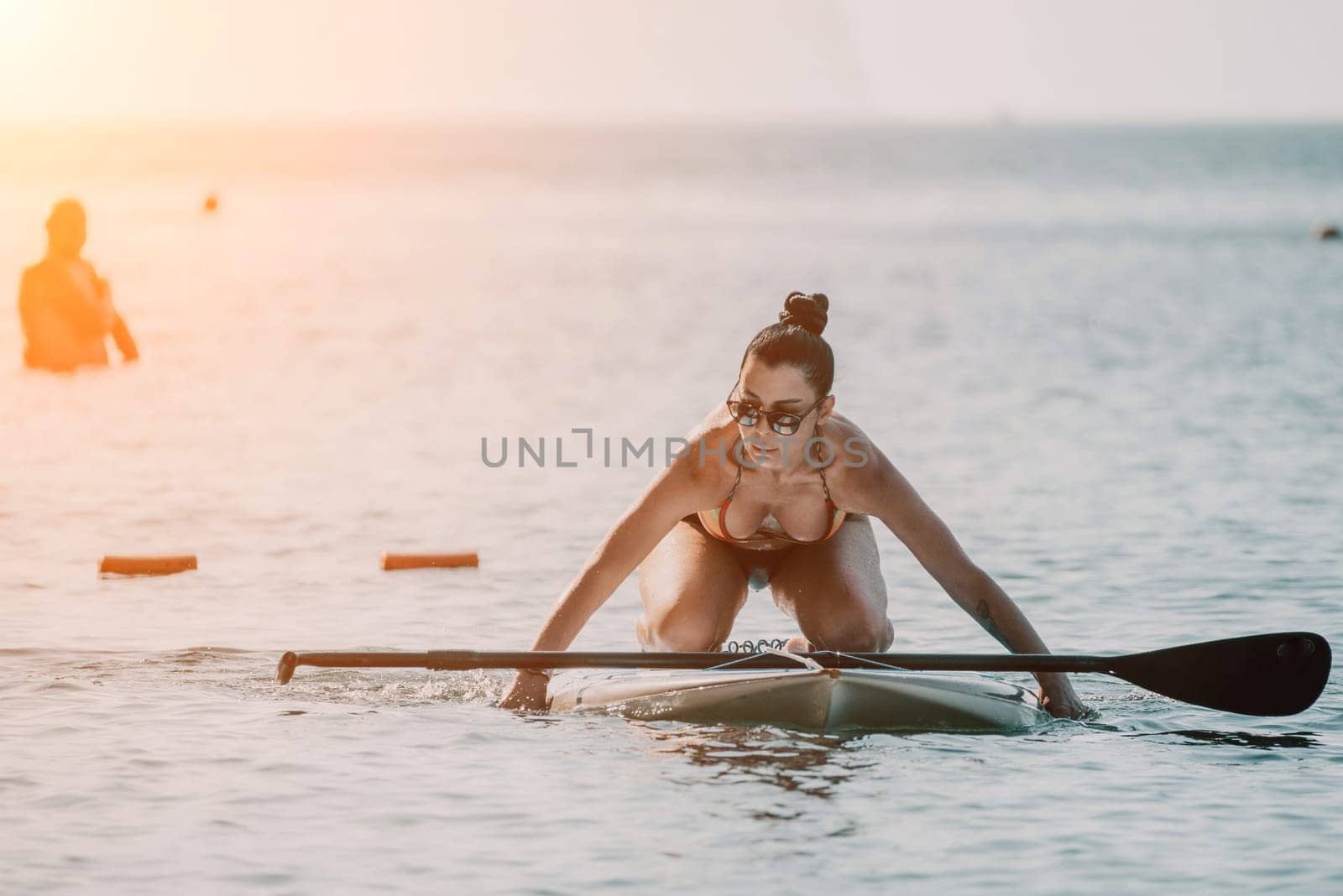 Sea woman sup. Silhouette of happy middle aged tanned woman in rainbow bikini, surfing on SUP board, confident paddling through water surface. Idyllic sunset. Active lifestyle at sea or river. by panophotograph