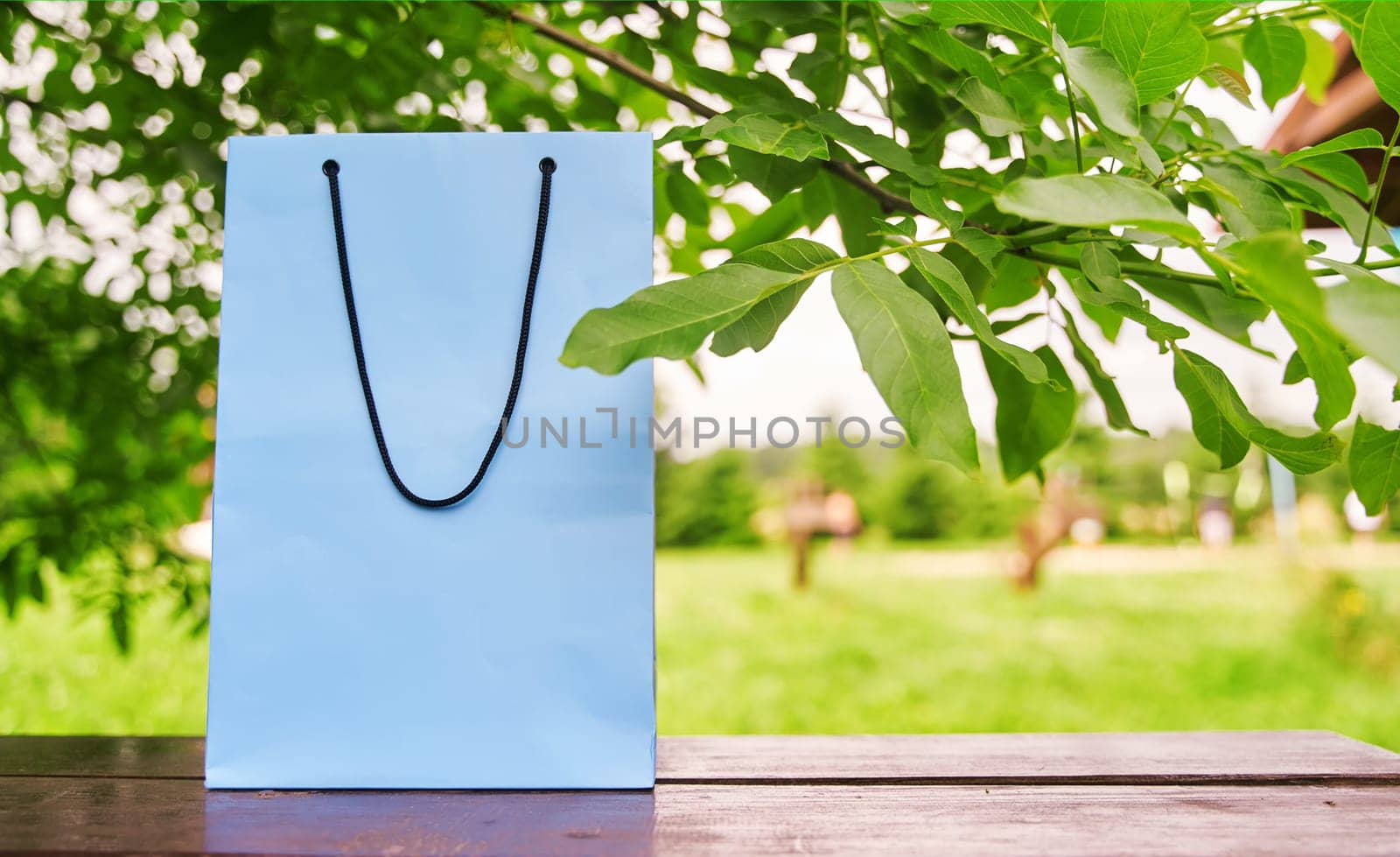 Blank paper bag stands on a wooden table against the background of green leaves. by driver-s