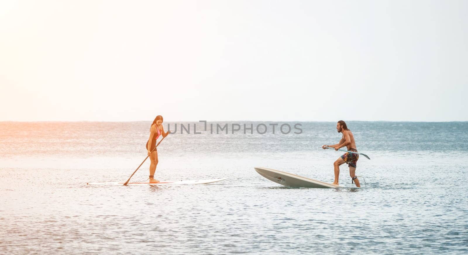 Sea woman and man on sup. Silhouette of happy young woman and man, surfing on SUP board, confident paddling through water surface. Idyllic sunset. Active lifestyle at sea or river. by panophotograph