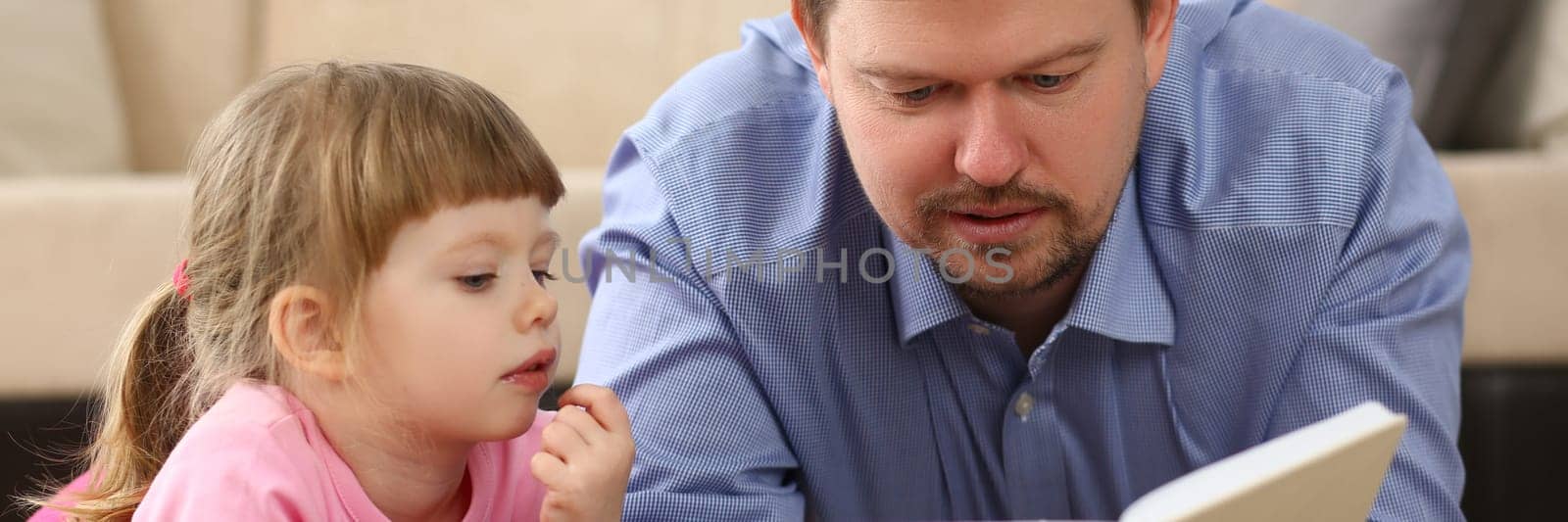 Loving dad lies on warm floor in living room with cute little daughter. Caring father enjoying fairy tale with happy child concept