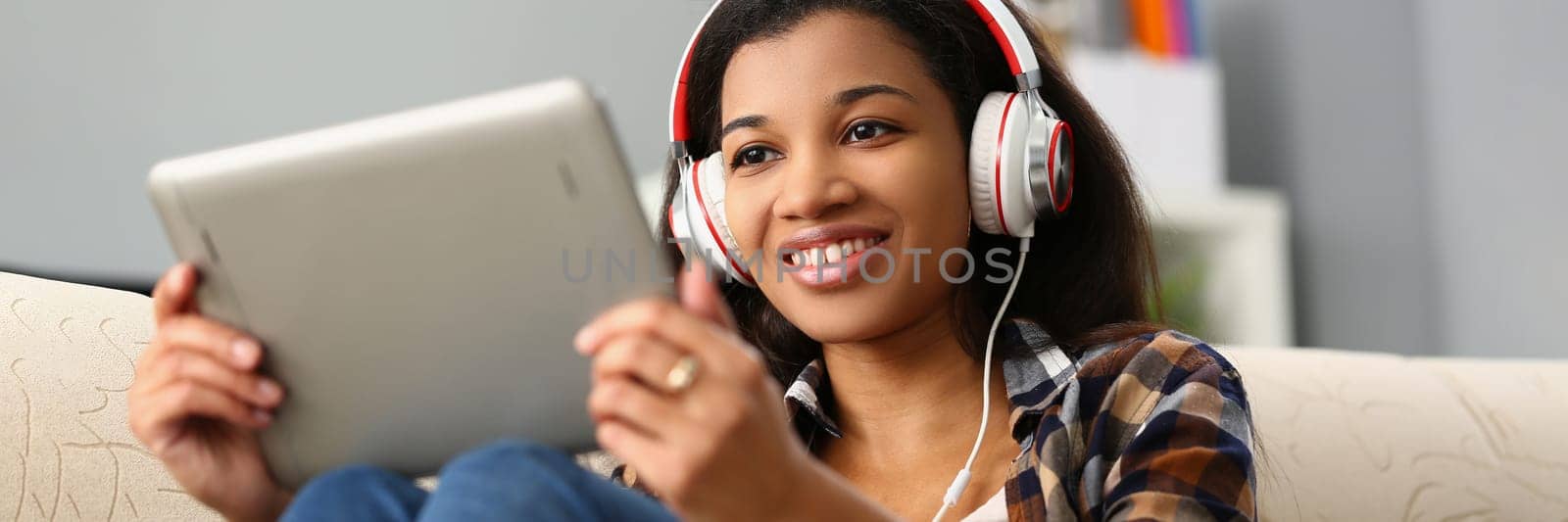 Relaxed charming african american woman in headphones listening to music and choosing with finger on tablet screen. Applications for watching movies concept