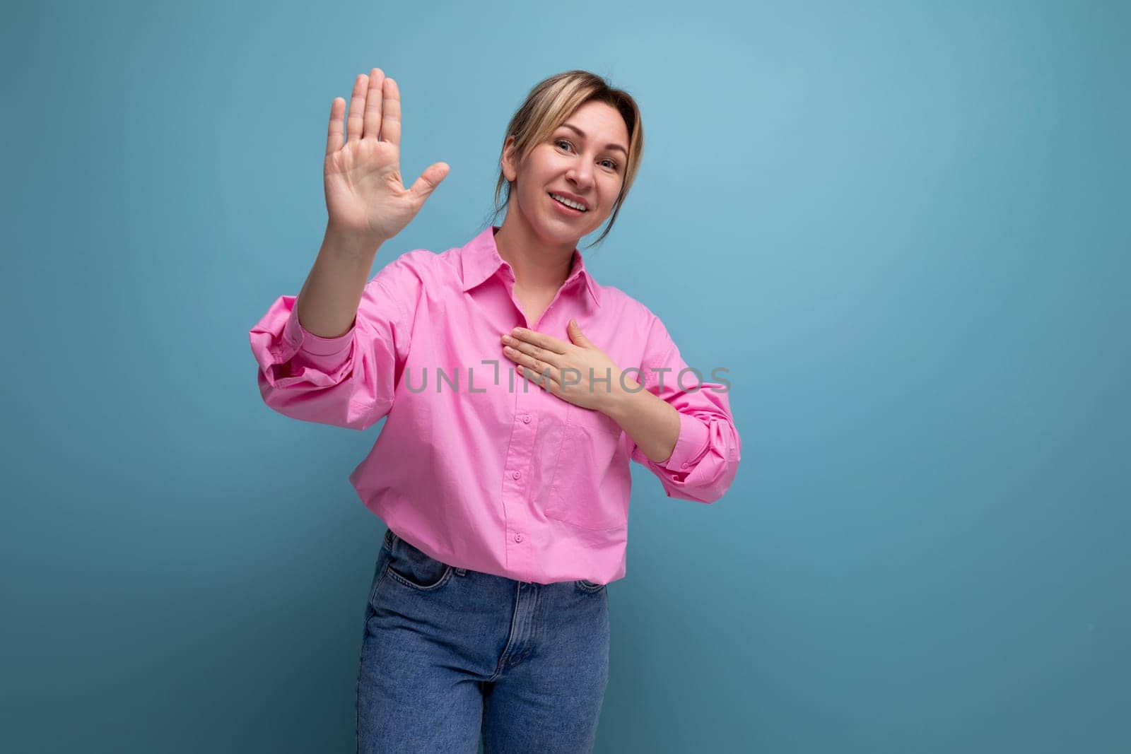 young well-groomed pretty european blonde secretary woman dressed in a pink shirt shows her hand to the side of an advertisement offer on a studio background with copy space.