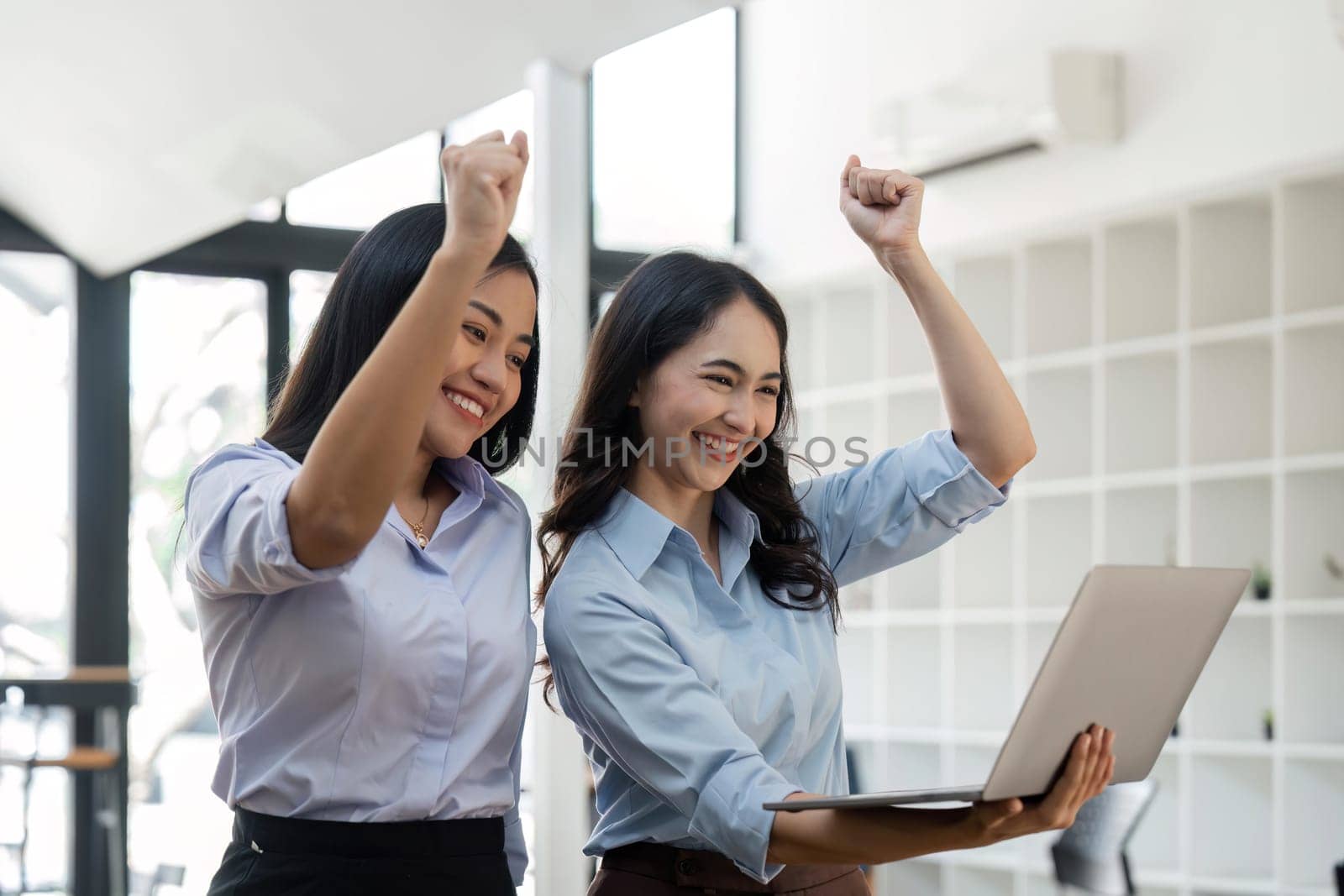 Two young businesswomen show joyful expression of success at work smiling happily with a laptop computer in a modern office.