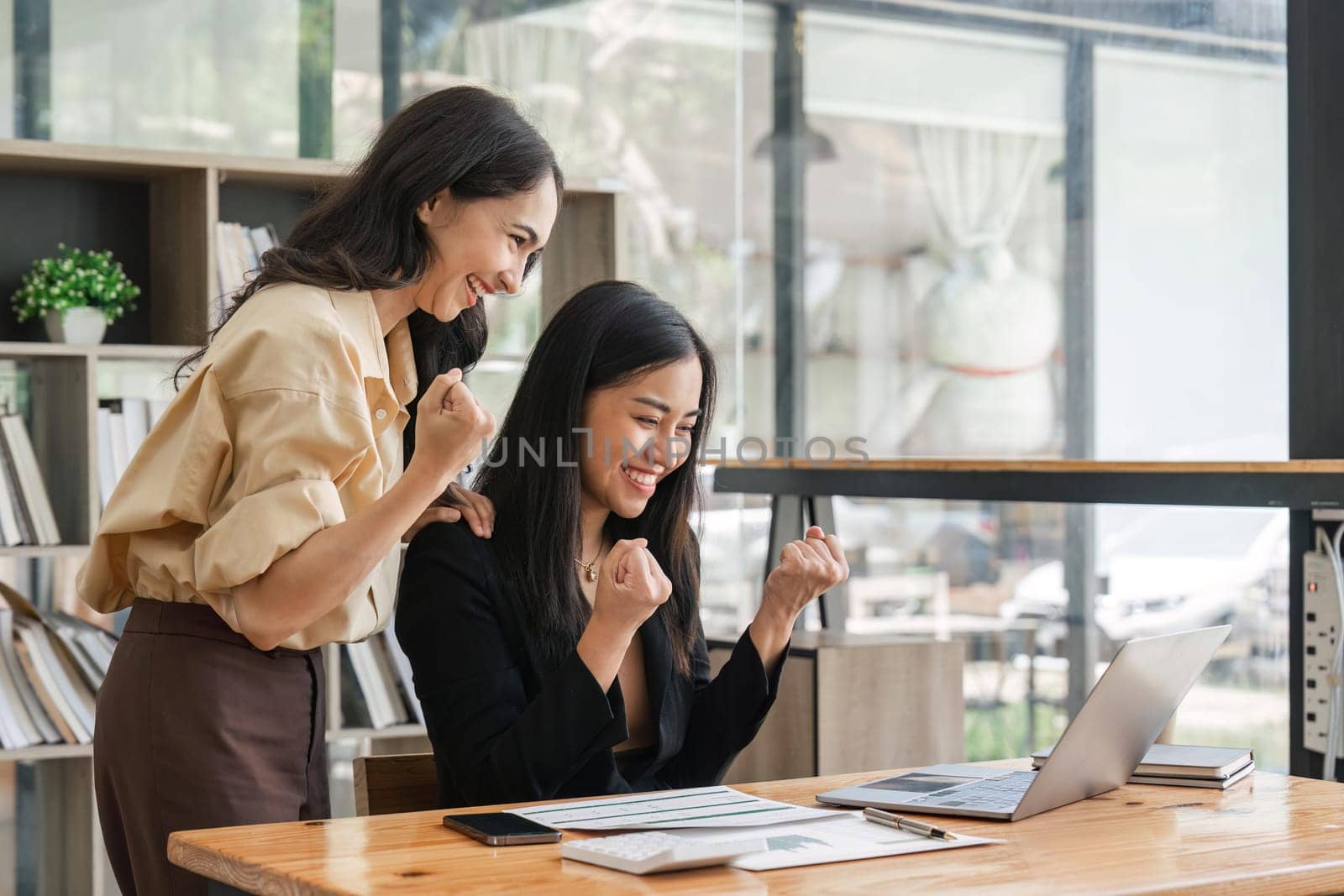 Two young businesswomen show joyful expression of success at work smiling happily with a laptop computer in a modern office.