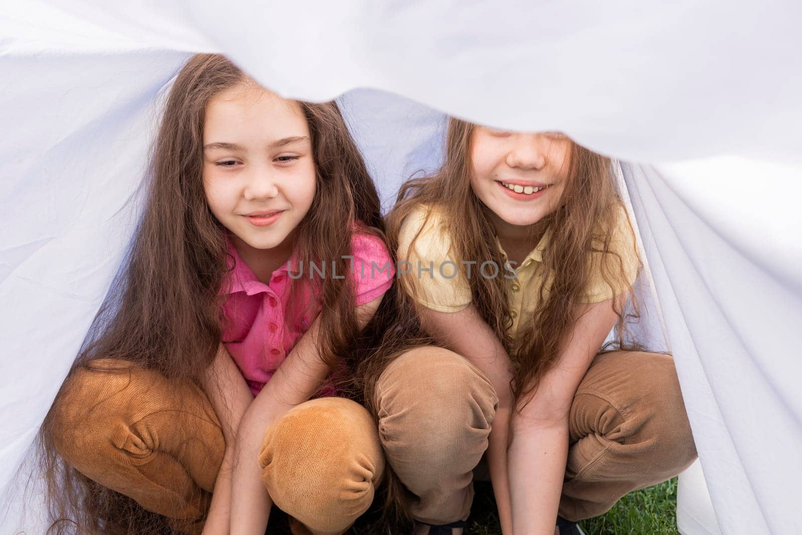 Two Little Girls, Sisters With Long Hair Sits Under Sheet On Grass in Meadow. Caucasian Asian Siblings Having Fun, Joy Together. Family Love And Care, True Friendship. Horizontal Plane. by netatsi
