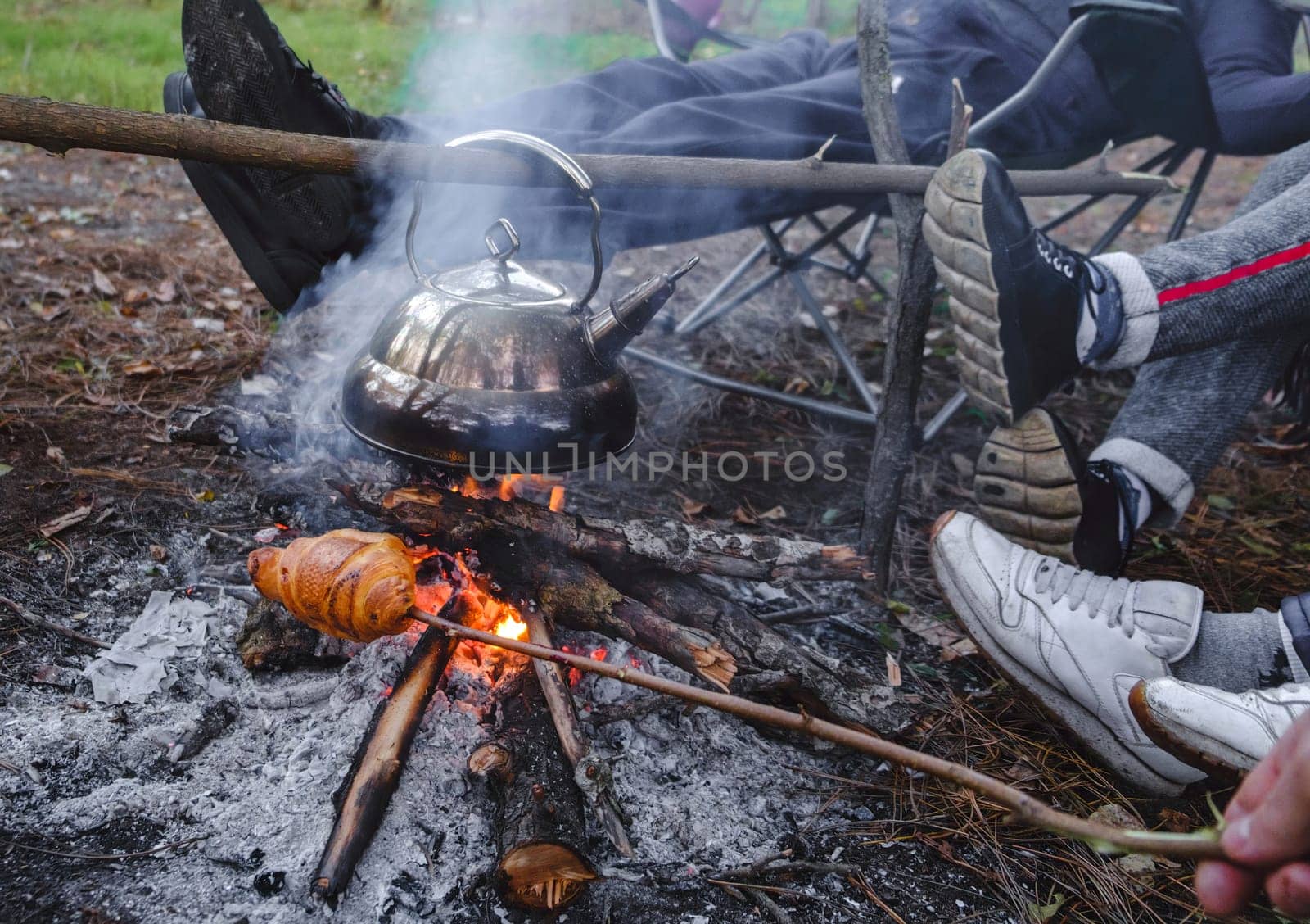 Young and cheerful friends sitting at bonfire , boil the kettle and fry bread on the nature. cooking on the campfire by igor010