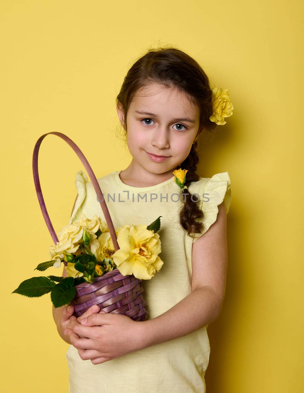 Beauty portrait Caucasian noble charming little girl in yellow elegant wear and flowers in hair, holding bouquet of yellow roses in purple wicker basket, smiling looking at camera, isolated background