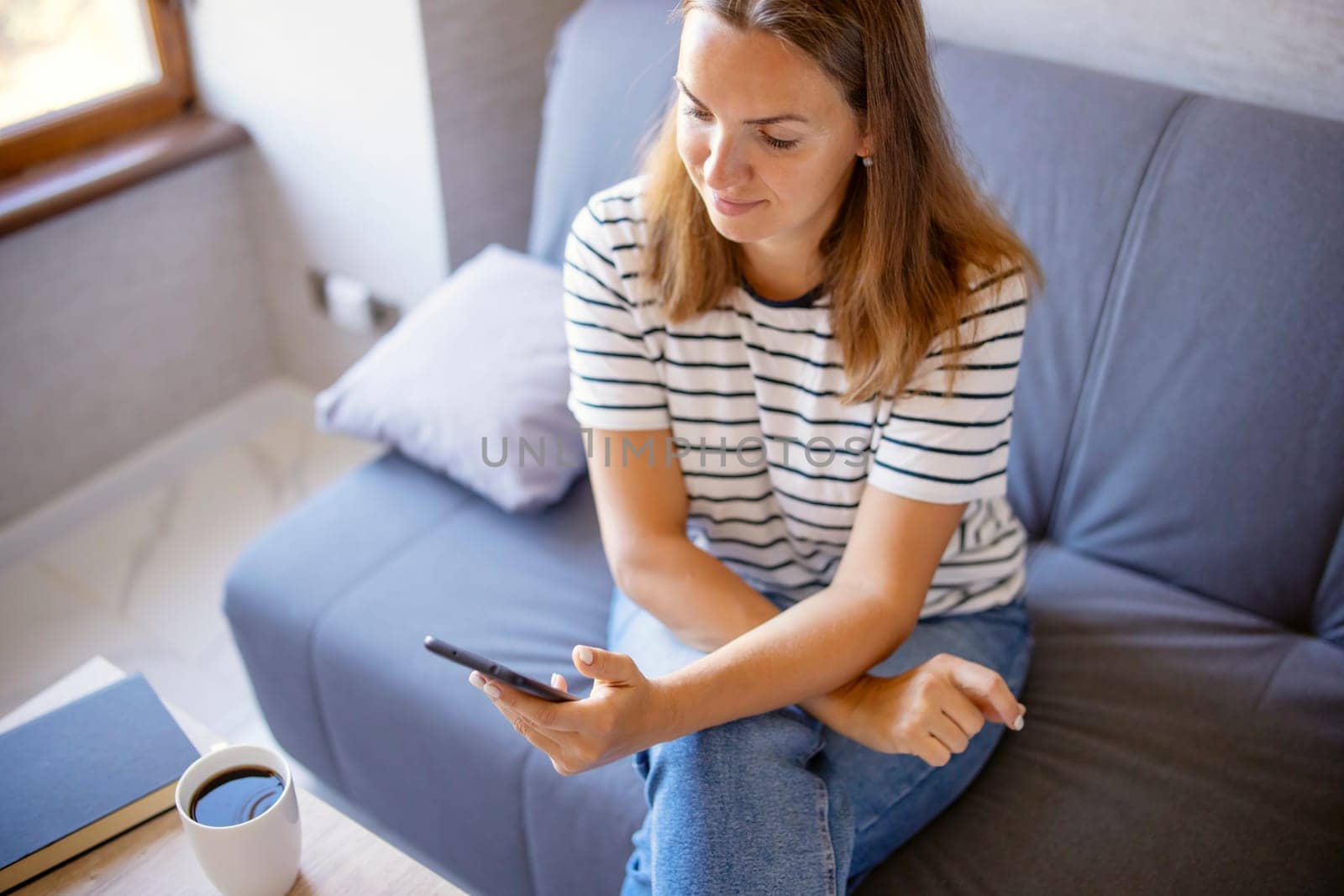 Young woman sitting, relaxing on sofa in living room, browsing wireless internet on smartphone, text message on modern mobile phone, shopping online through website.