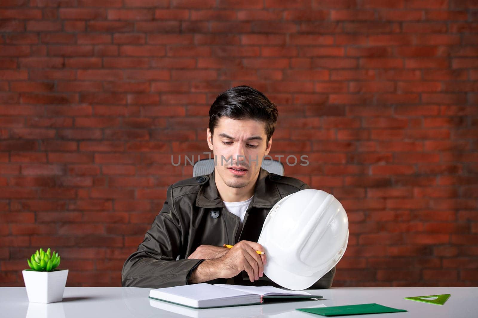 front view bored male engineer sitting behind his working place and writing notes document plan business corporate property builder contractor job agenda by Kamran