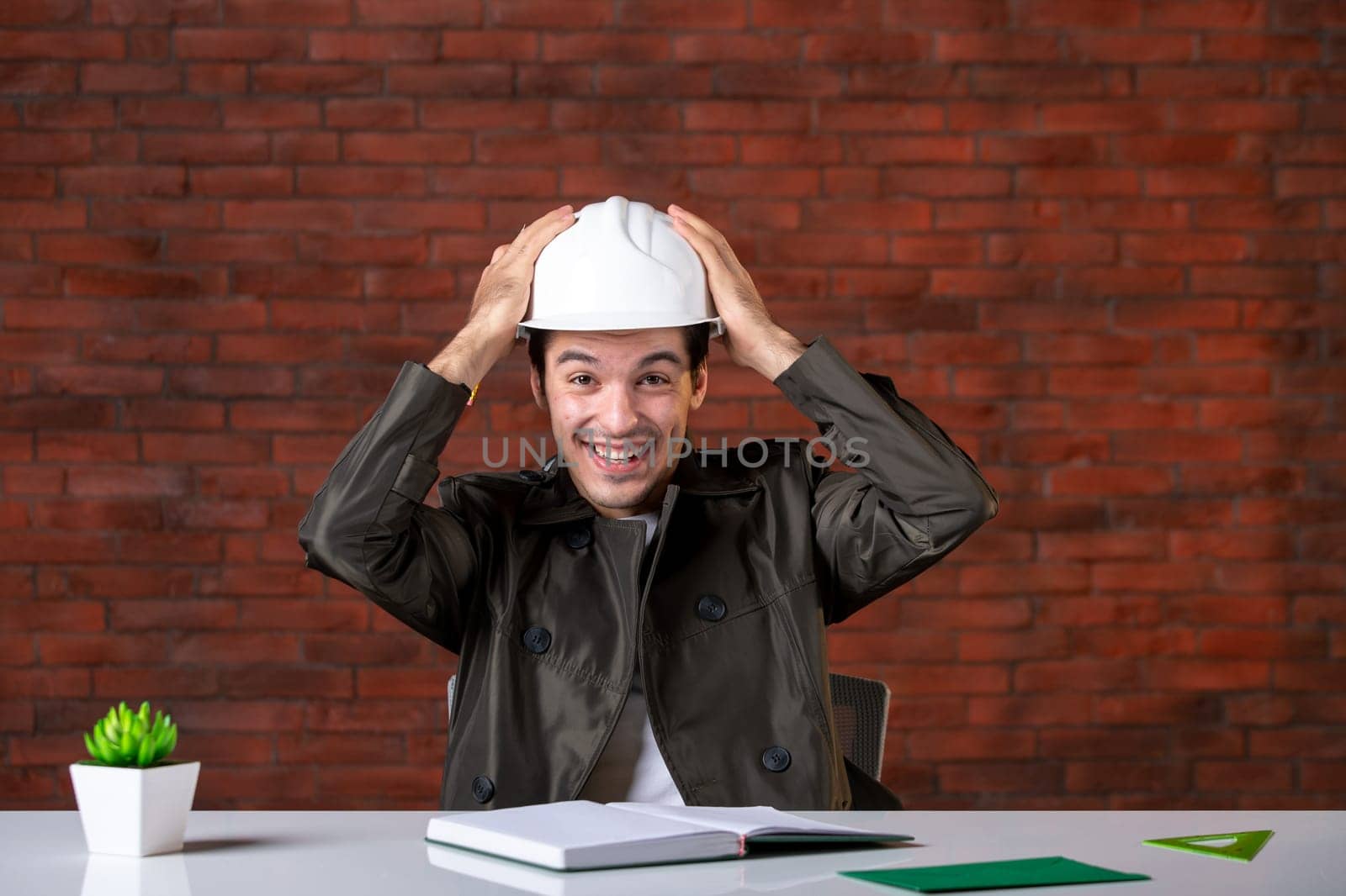 front view happy male engineer sitting behind his working place and writing notes document plan business contractor agenda builder corporate job property by Kamran