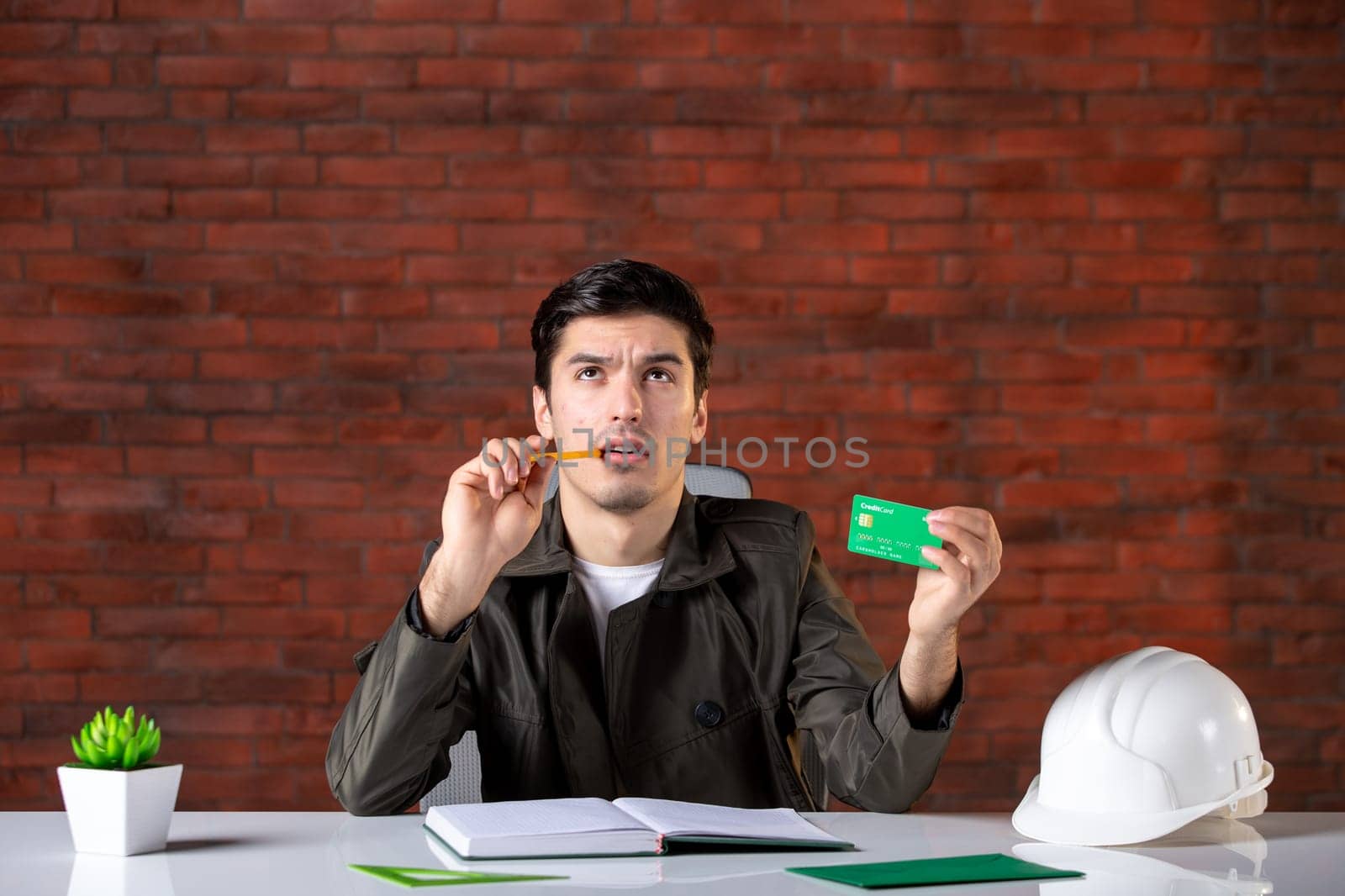 front view male engineer sitting behind his working place and holding green credit card builder money plan contractor business property agenda