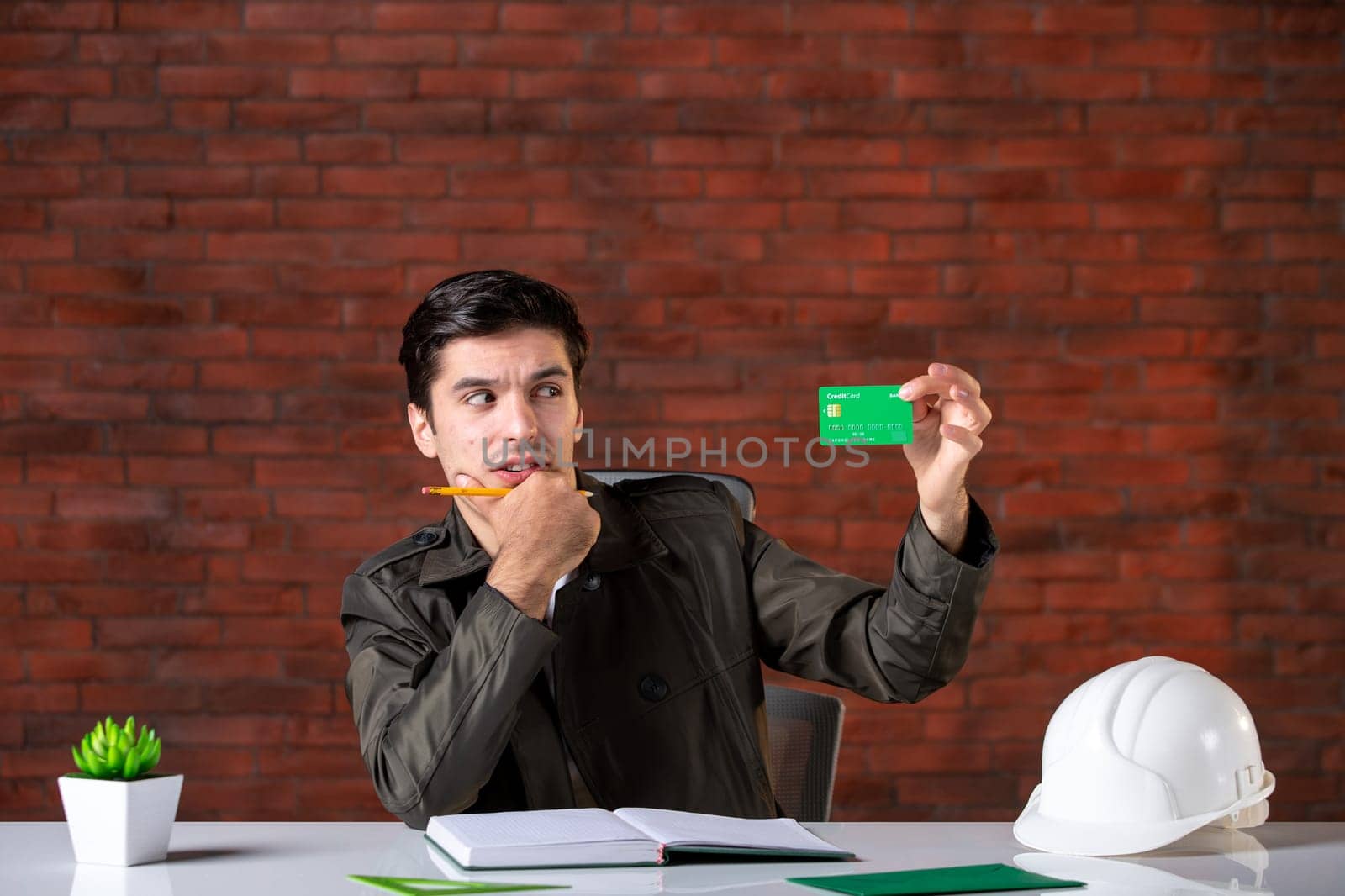 front view male engineer sitting behind his working place and holding green credit card builder money plan corporate contractor business agenda