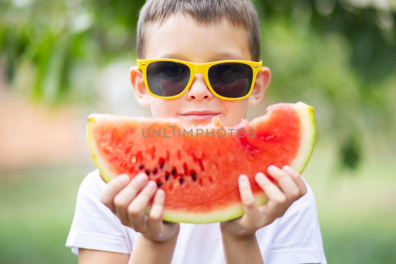 Cute boy in yellow sunglasses eating watermelon outdoors in summer. Healthy eating seasonal berries and fruits.