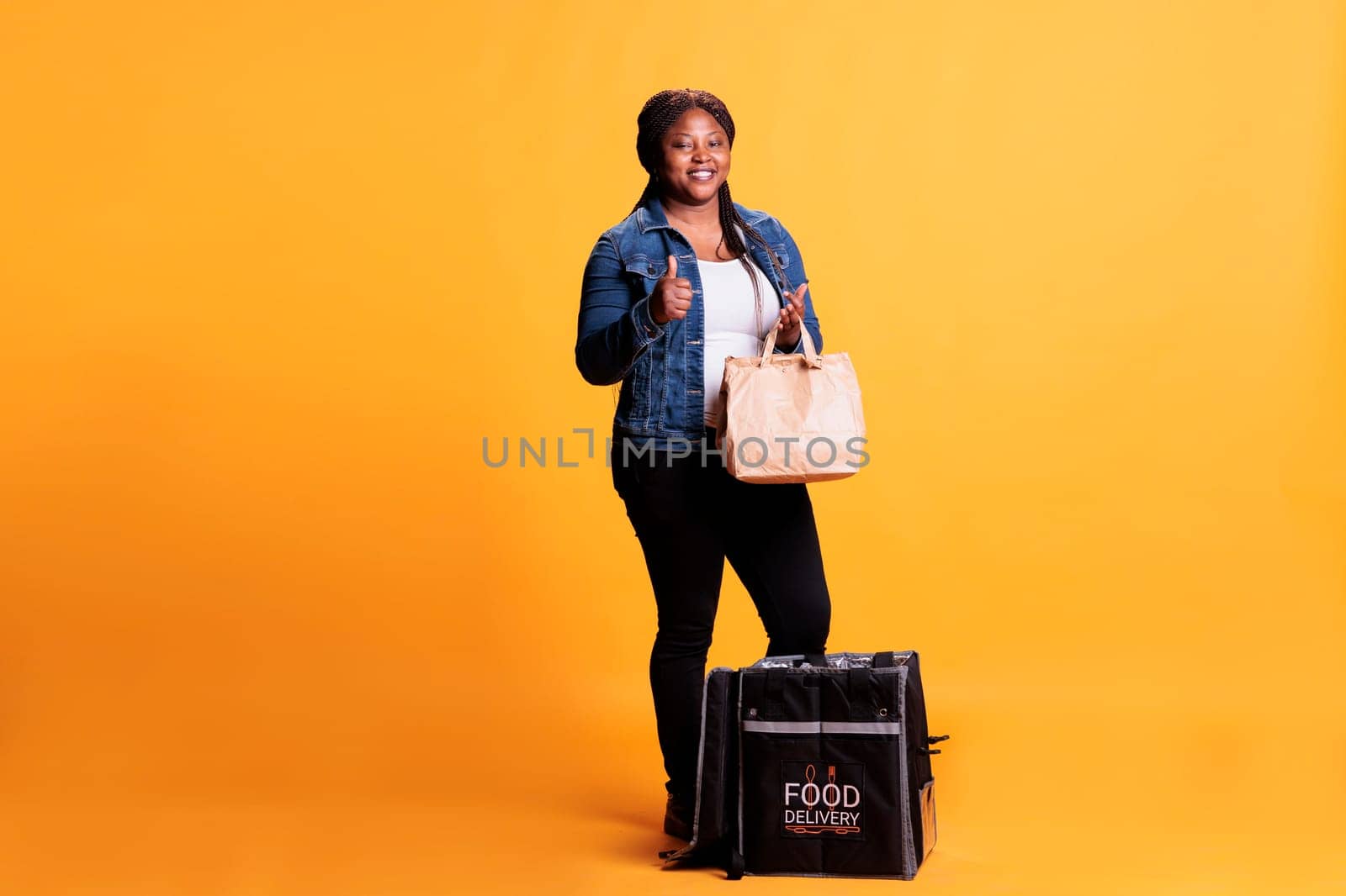 Full body of restaurant worker holding paper bag full with takeaway order while doing thumb up sign at camera approving delivery. Food transportation service and take away concept