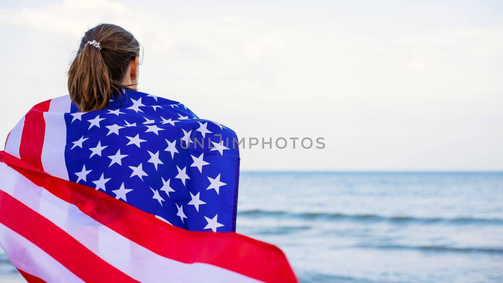 Back view of young woman holding national American flag on a ocean beach. USA Memorial day and Independence day concept.