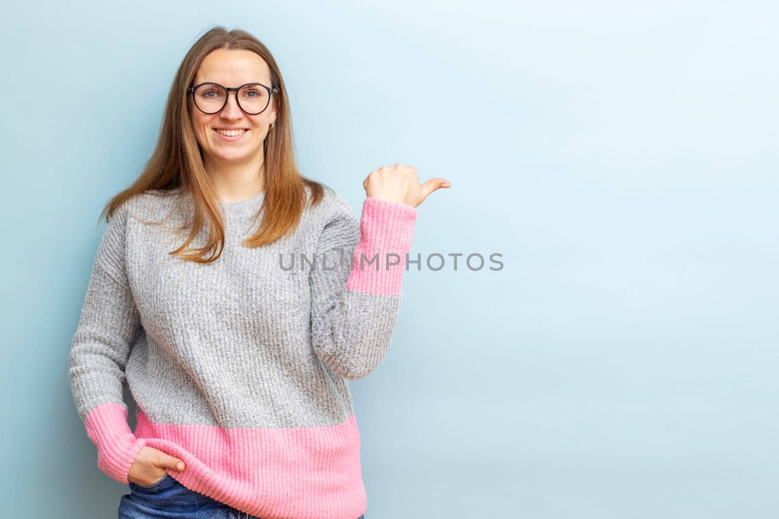 Young beautiful brunette woman in glasses smiling and pointing her finger to the side on blue background.