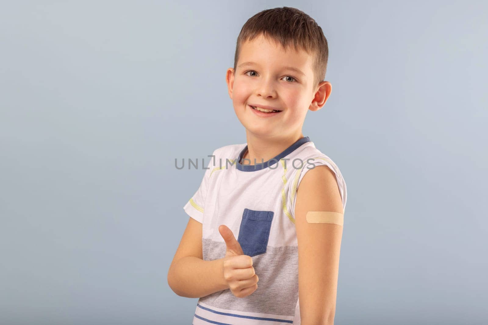 Vaccinated boy gesturing with thumbs up, showing hand with plaster cast after injection of coronavirus vaccine. Start of vaccination campaign for children and teenagers. Successful Covid-19 vaccination.