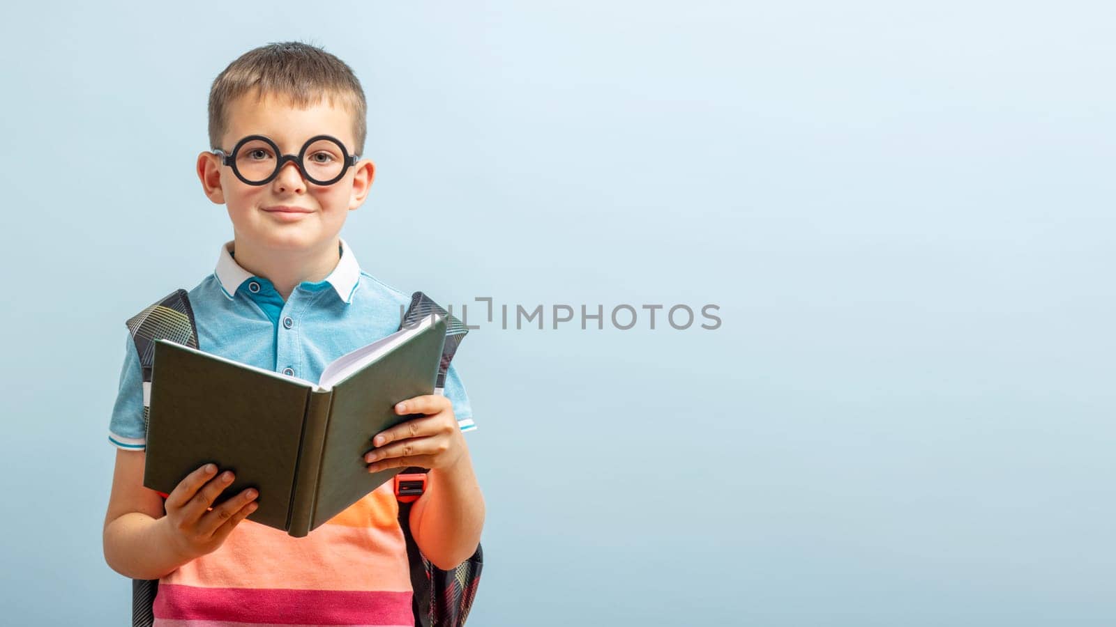 Little schoolboy in eyeglasses is reading a book on blue background. Education and school concept. Mockup, copy space